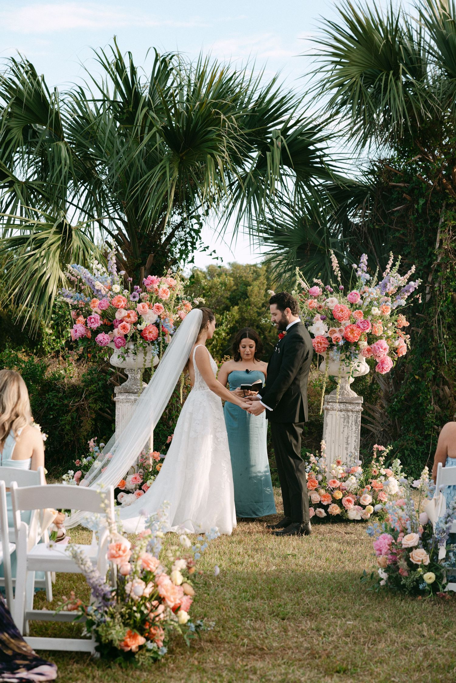 Bride and groom exchange vows at elegant outdoor ceremony with colorful floral urn arrangements.