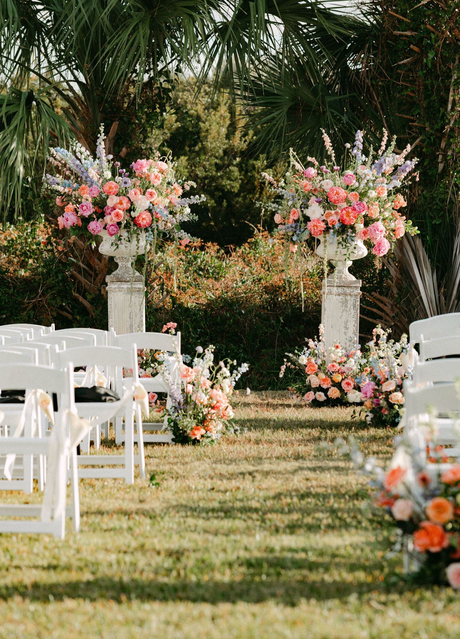 Elegant outdoor wedding ceremony aisle with white chairs and ornate stone urns filled with pink floral arrangements.