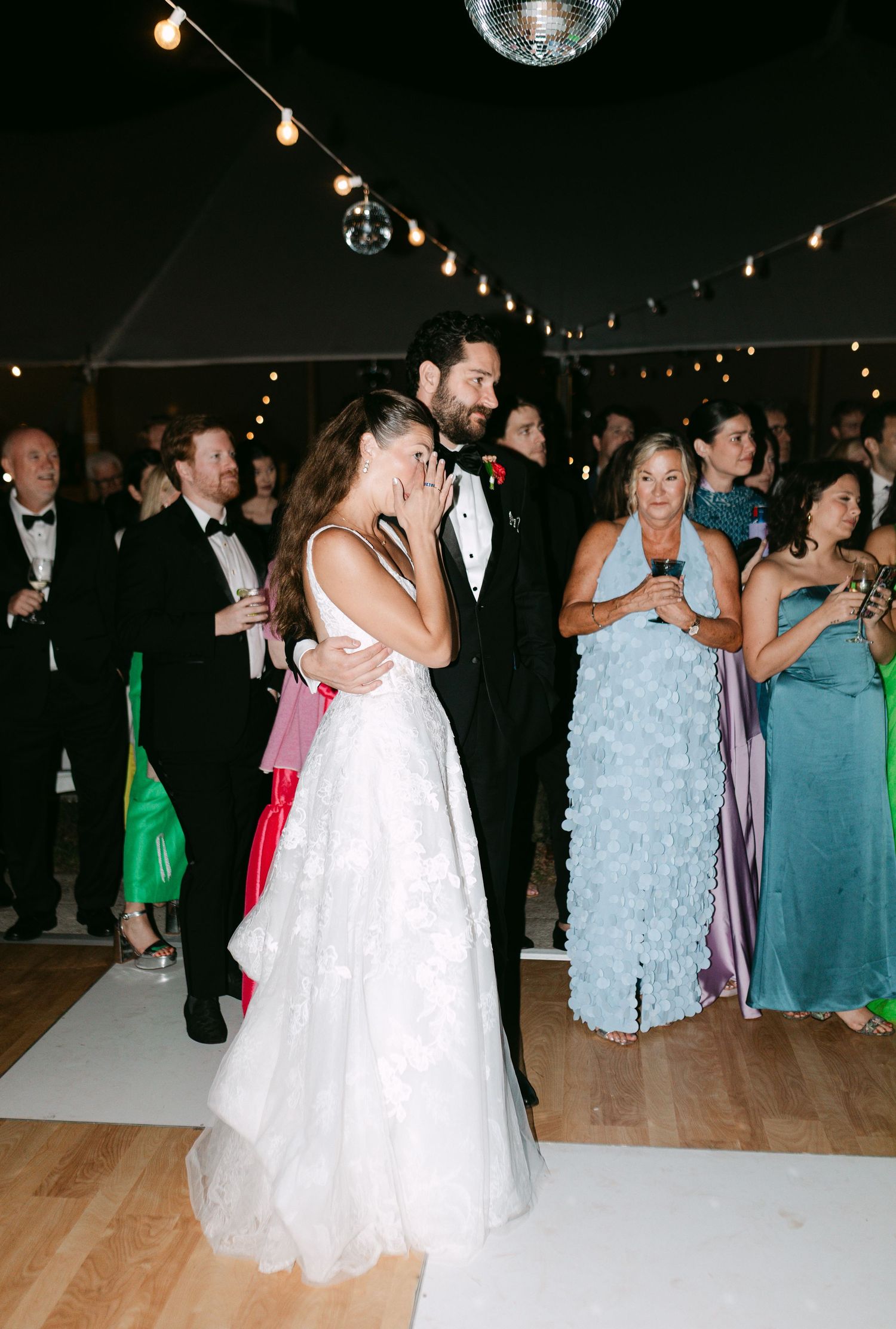 Newlyweds share emotional first dance embrace under disco ball while wedding guests watch at evening reception.