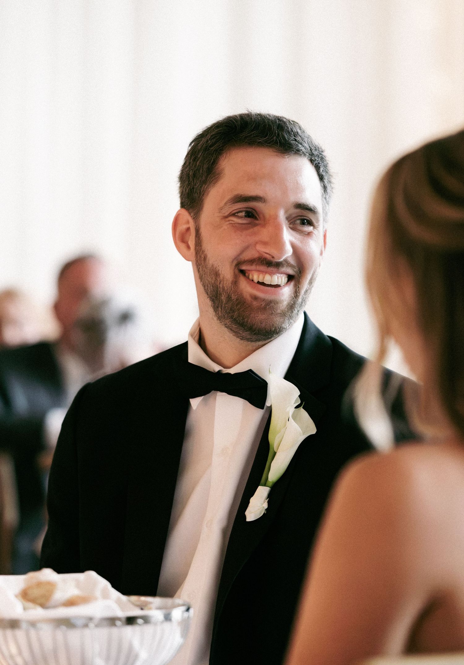 Smiling groom in black tuxedo with white boutonniere at elegant wedding reception dinner table.