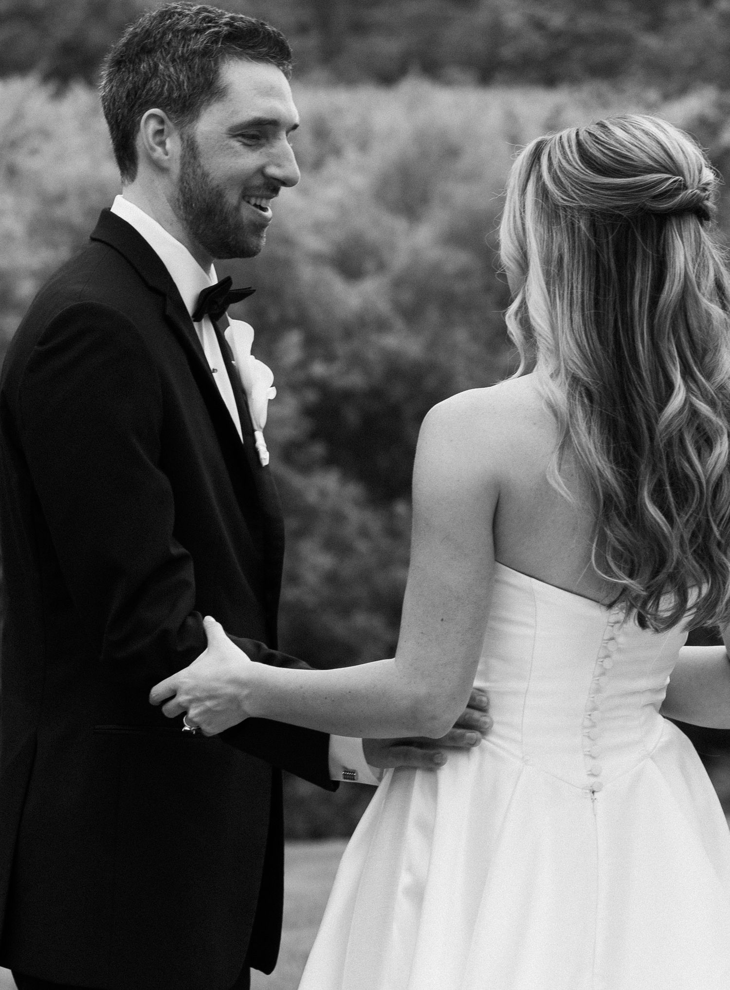 Black and white photo of smiling groom in tuxedo facing bride in strapless wedding gown holding hands outdoors.
