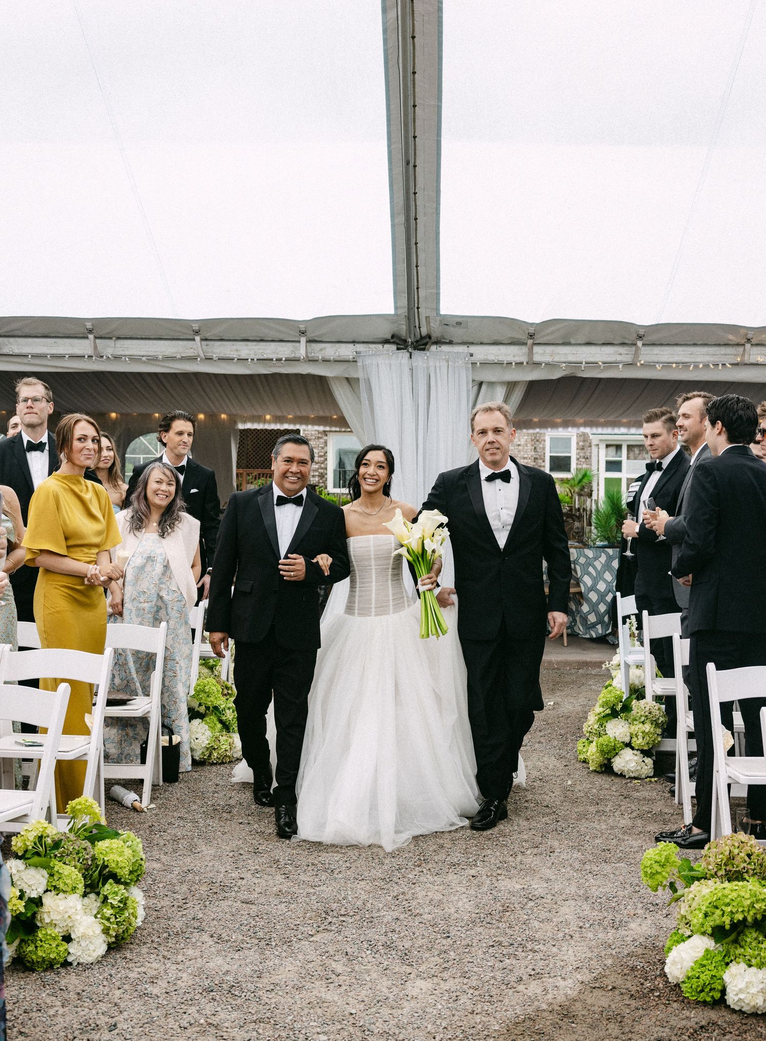 Bride walks down aisle with two escorts in tuxedos, flanked by white chairs and green hydrangeas under a tent.