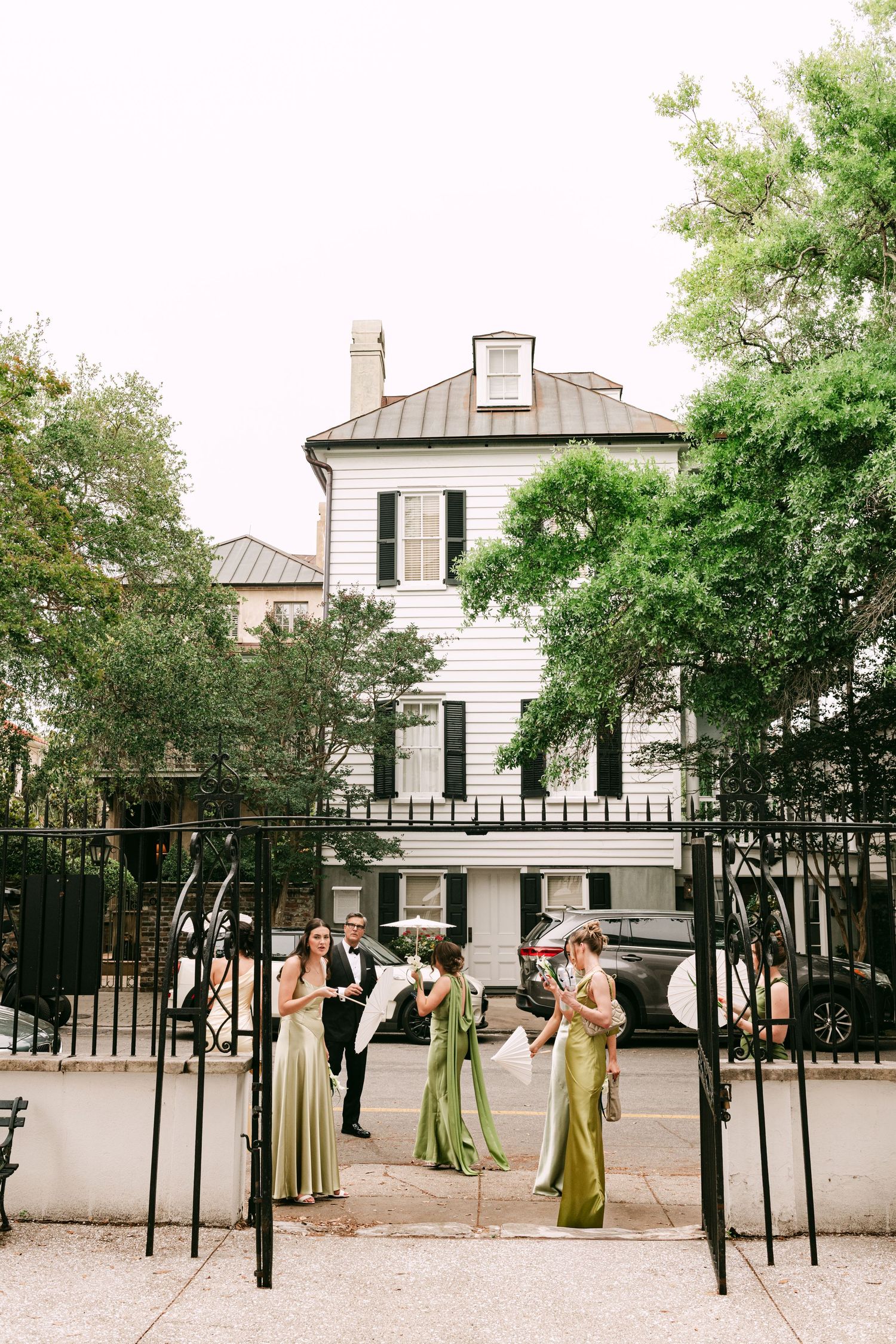 Bridesmaids in olive green gowns stand at black iron gates outside a white historic wedding venue.