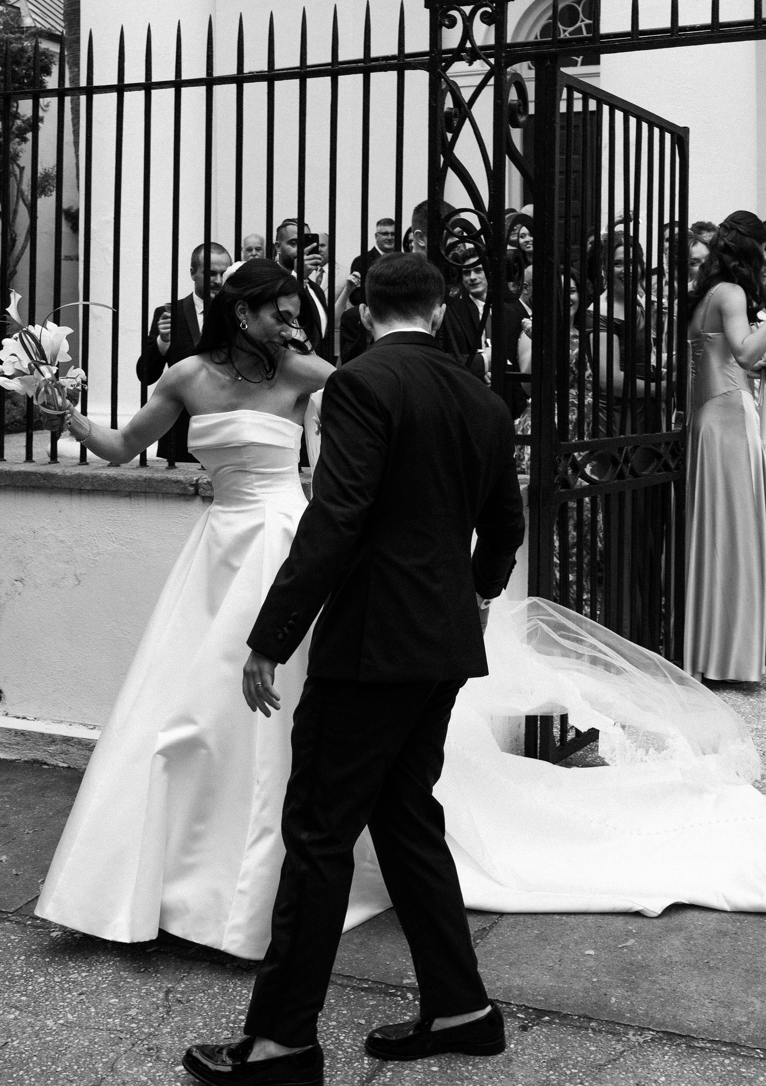 Bride and groom walking through iron gate surrounded by wedding guests in black and white photo.