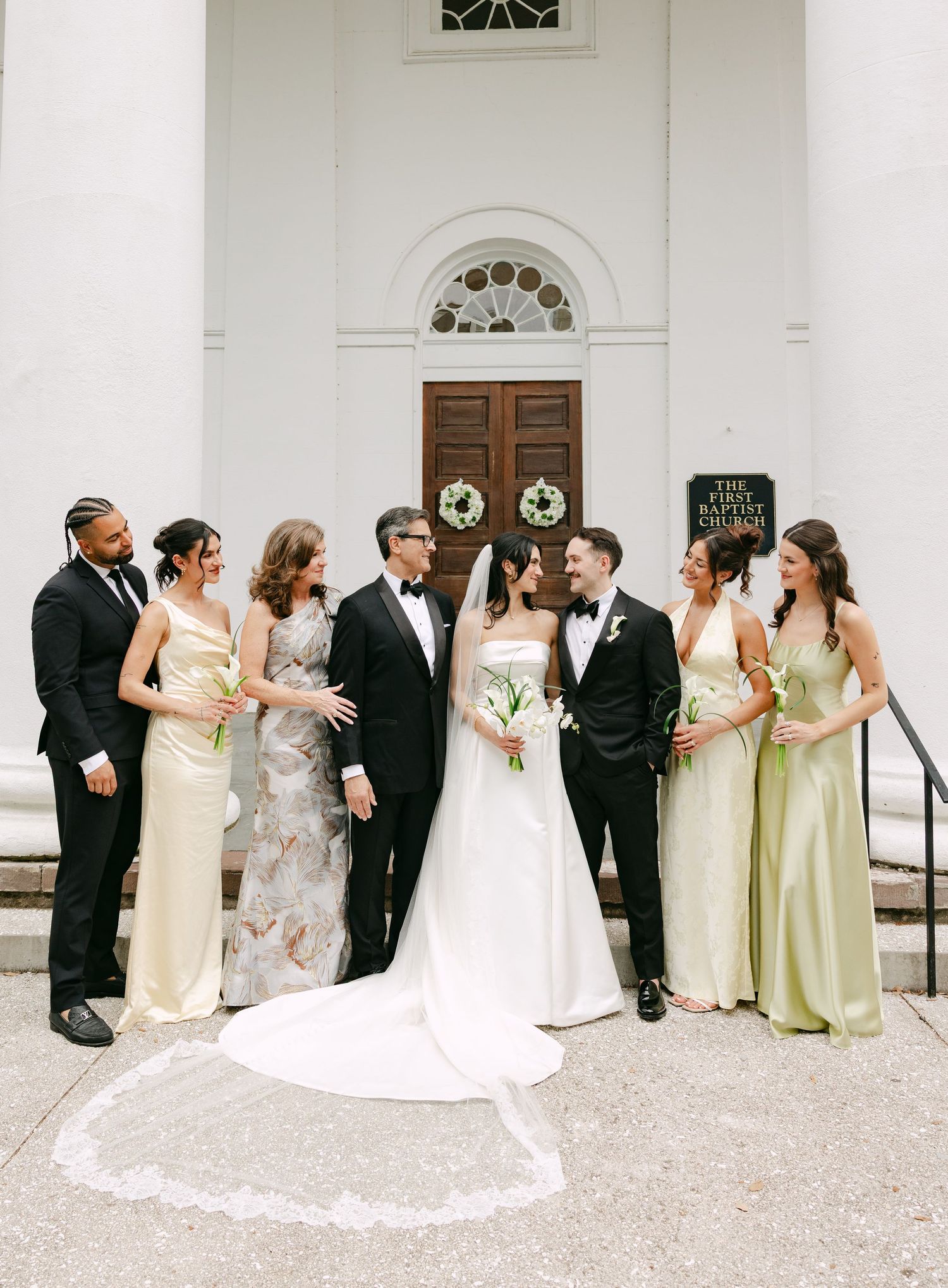 Wedding party in black tuxedos and sage green gowns gathered outside white church with wooden doors.