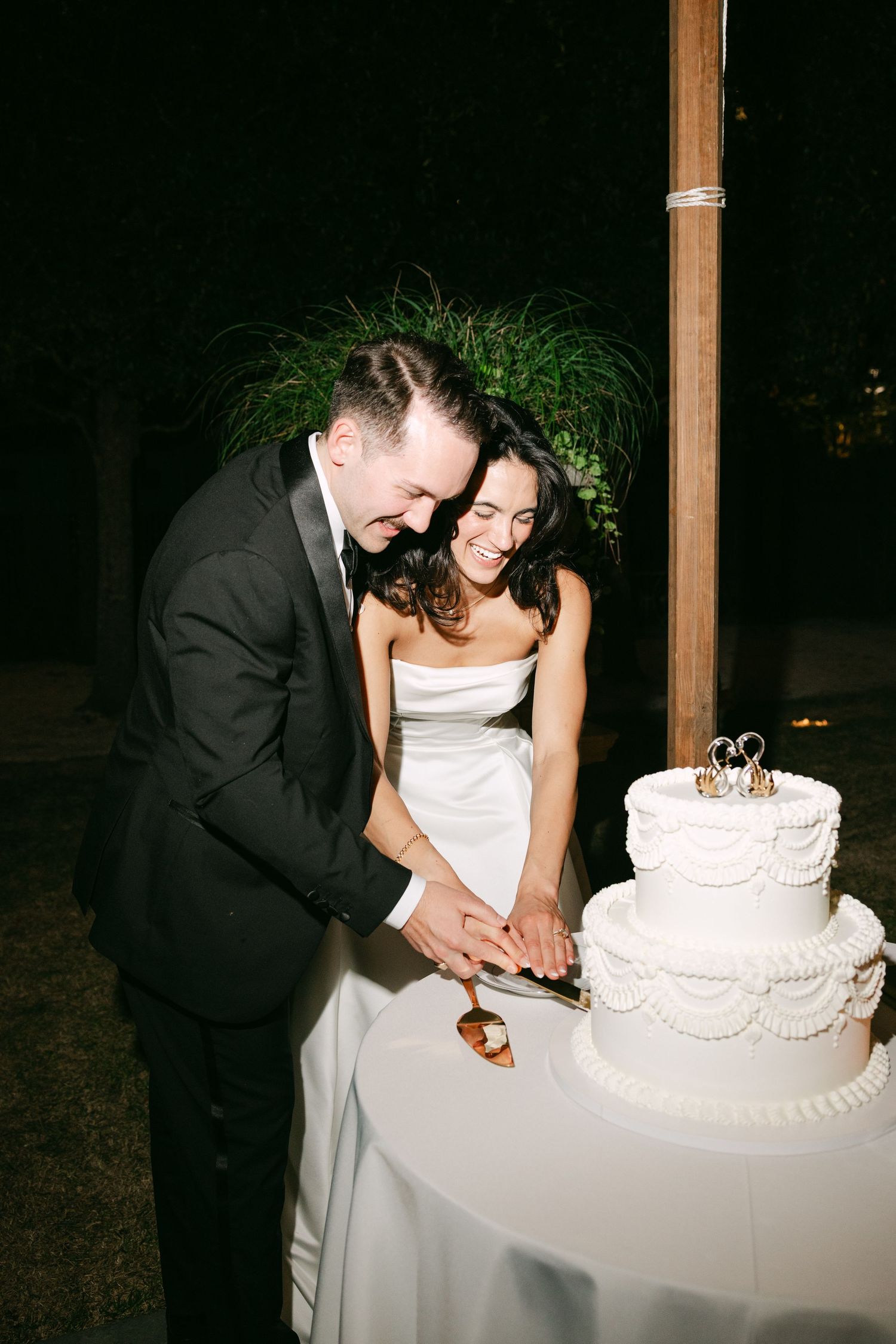 Newlyweds cut white tiered wedding cake together outdoors at night.