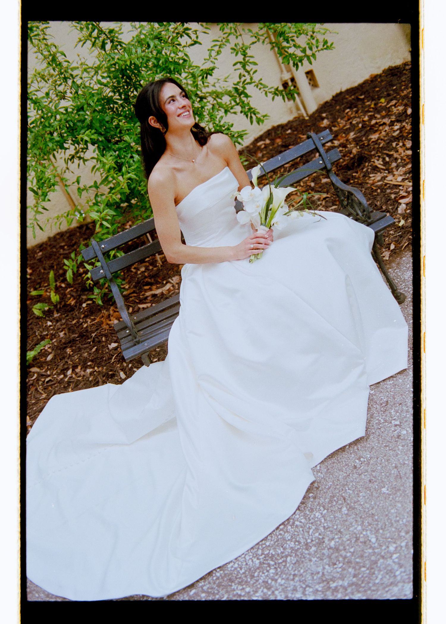 Elegant bride in strapless white gown with cathedral train sits on bench holding white calla lily bouquet.