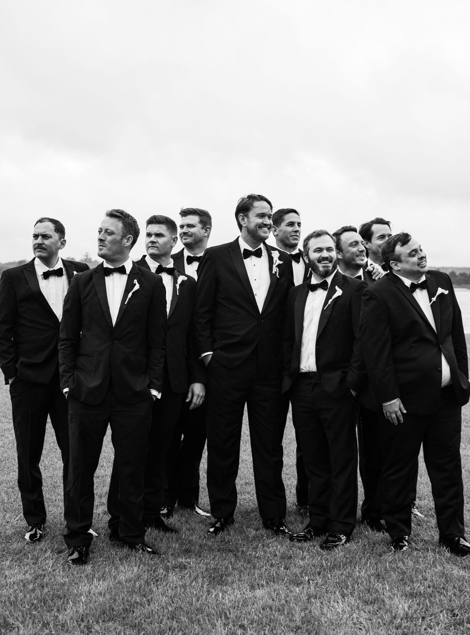 Groomsmen in black tuxedos with bow ties posing together outdoors on a cloudy wedding day.