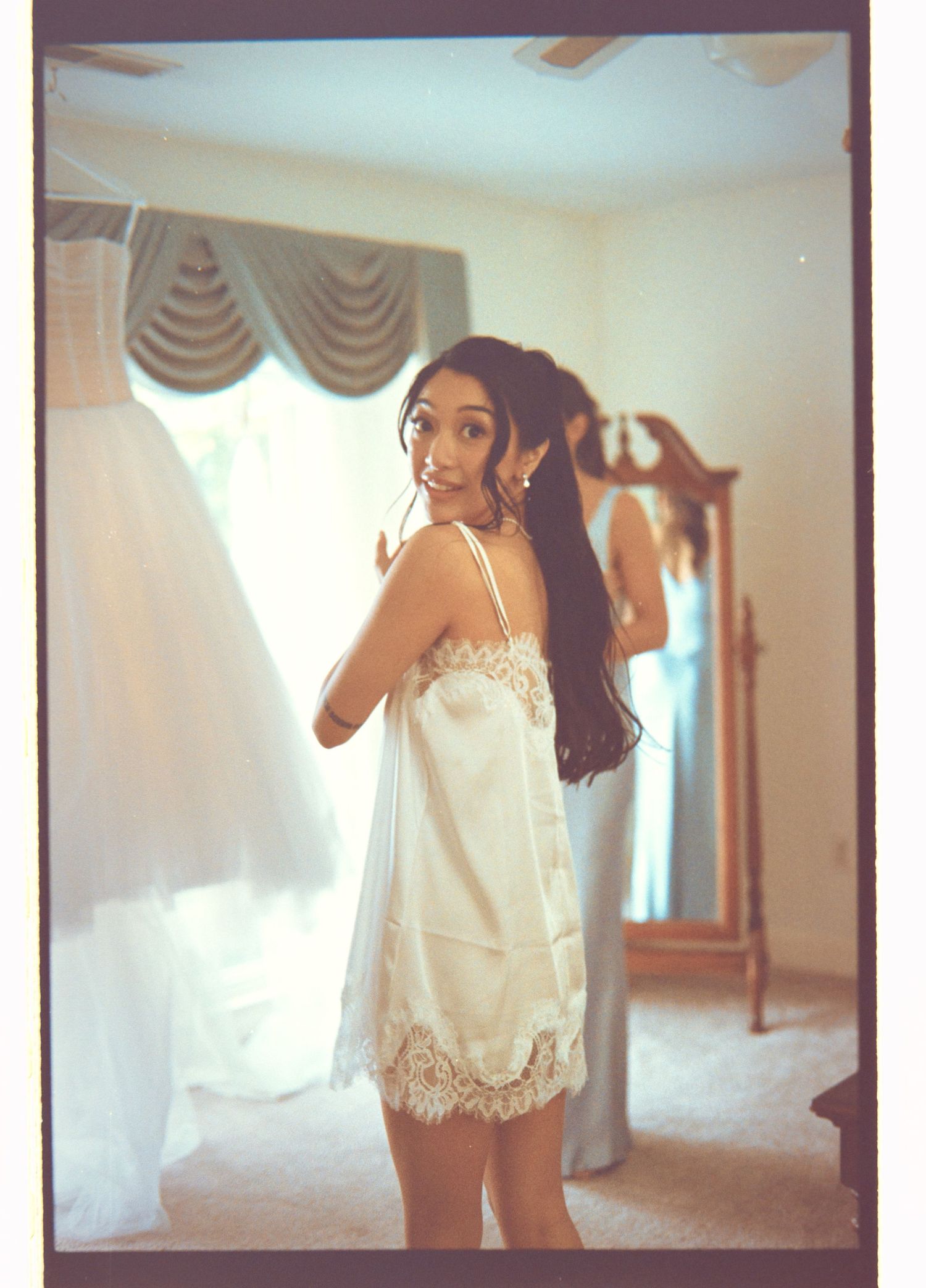 Bride in white lace slip dress smiling over shoulder in bedroom with wedding gown hanging nearby.