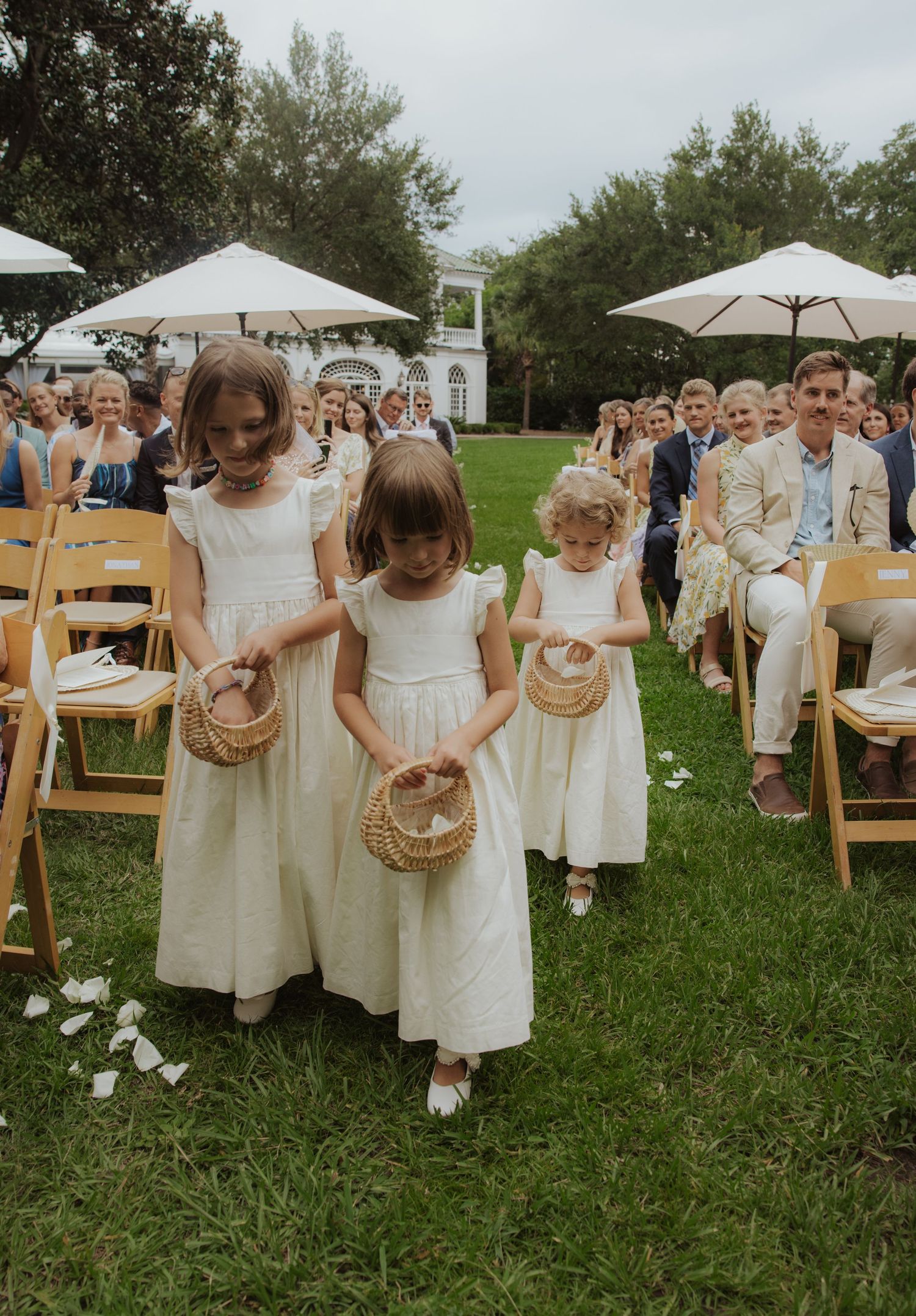 Young children in white dresses carry flower baskets while walking on grass near outdoor event seating and white umbrellas.