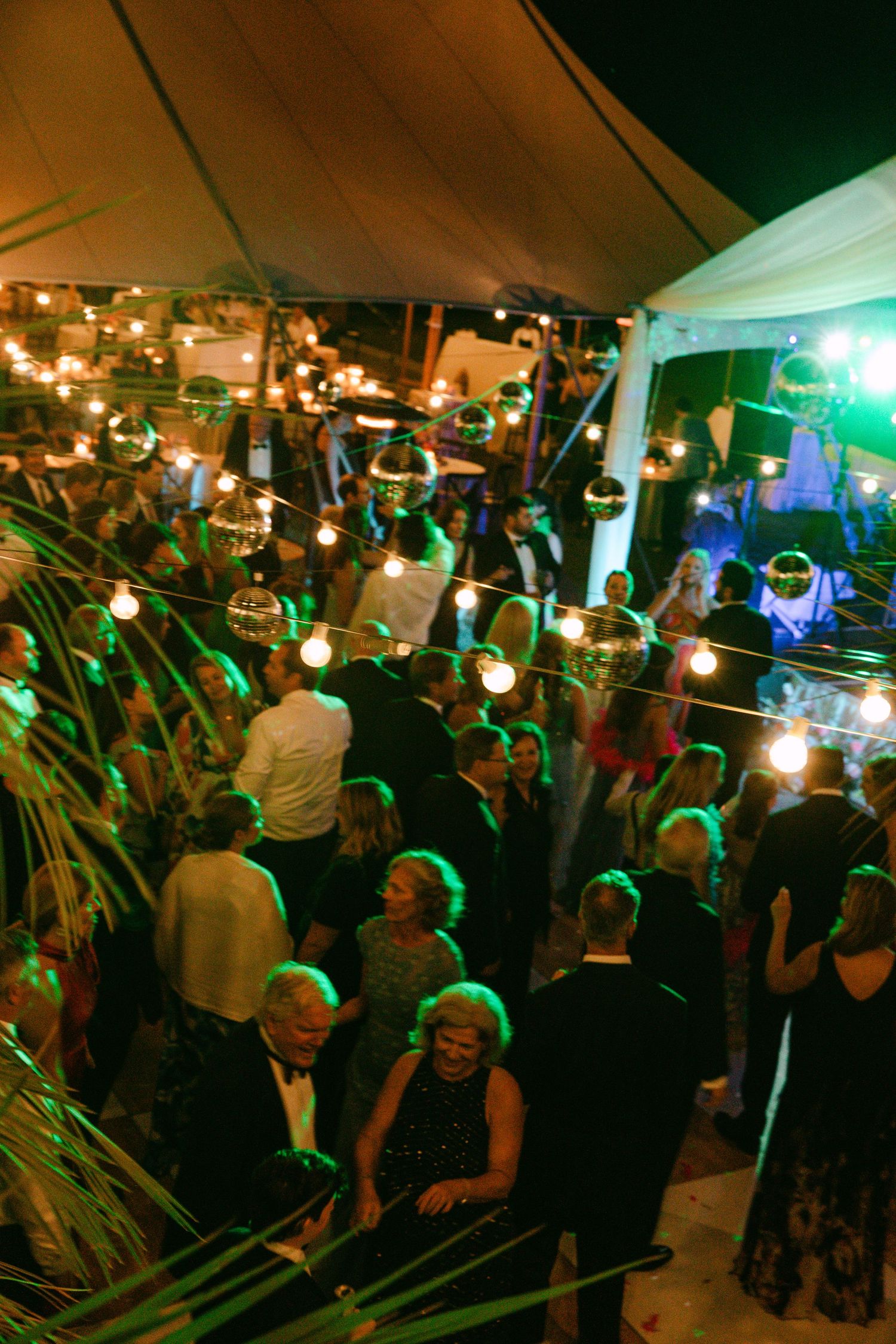 Guests dance under string lights and disco balls at lively outdoor tented wedding reception at night.