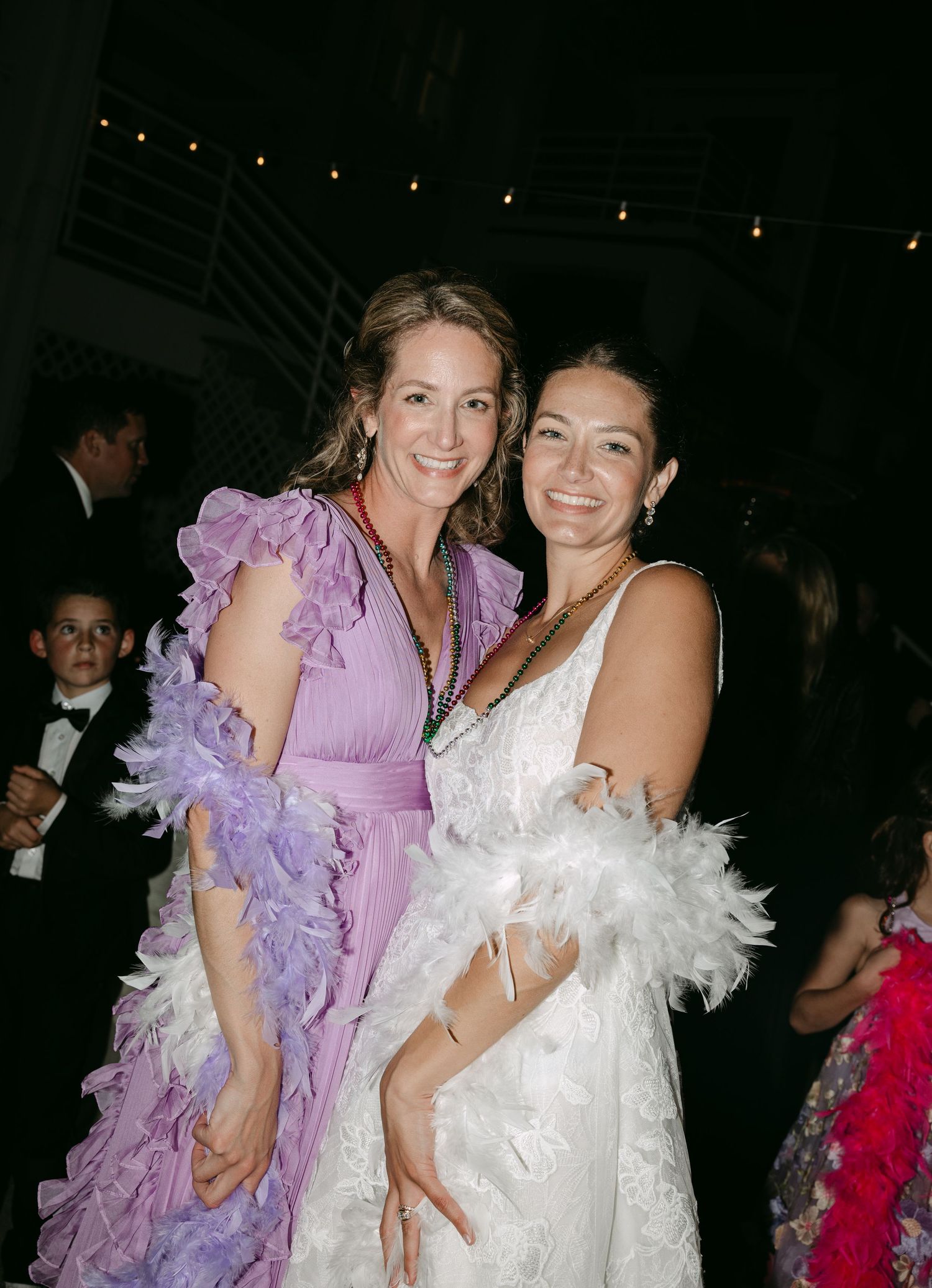 Two elegantly dressed women pose together at a formal event, wearing feather boas and festive gowns.