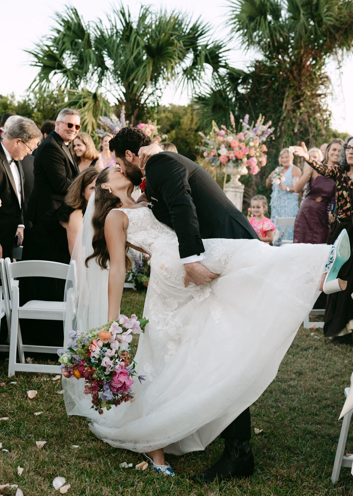 Groom dips bride for a kiss at outdoor wedding ceremony while guests celebrate with colorful floral bouquet.