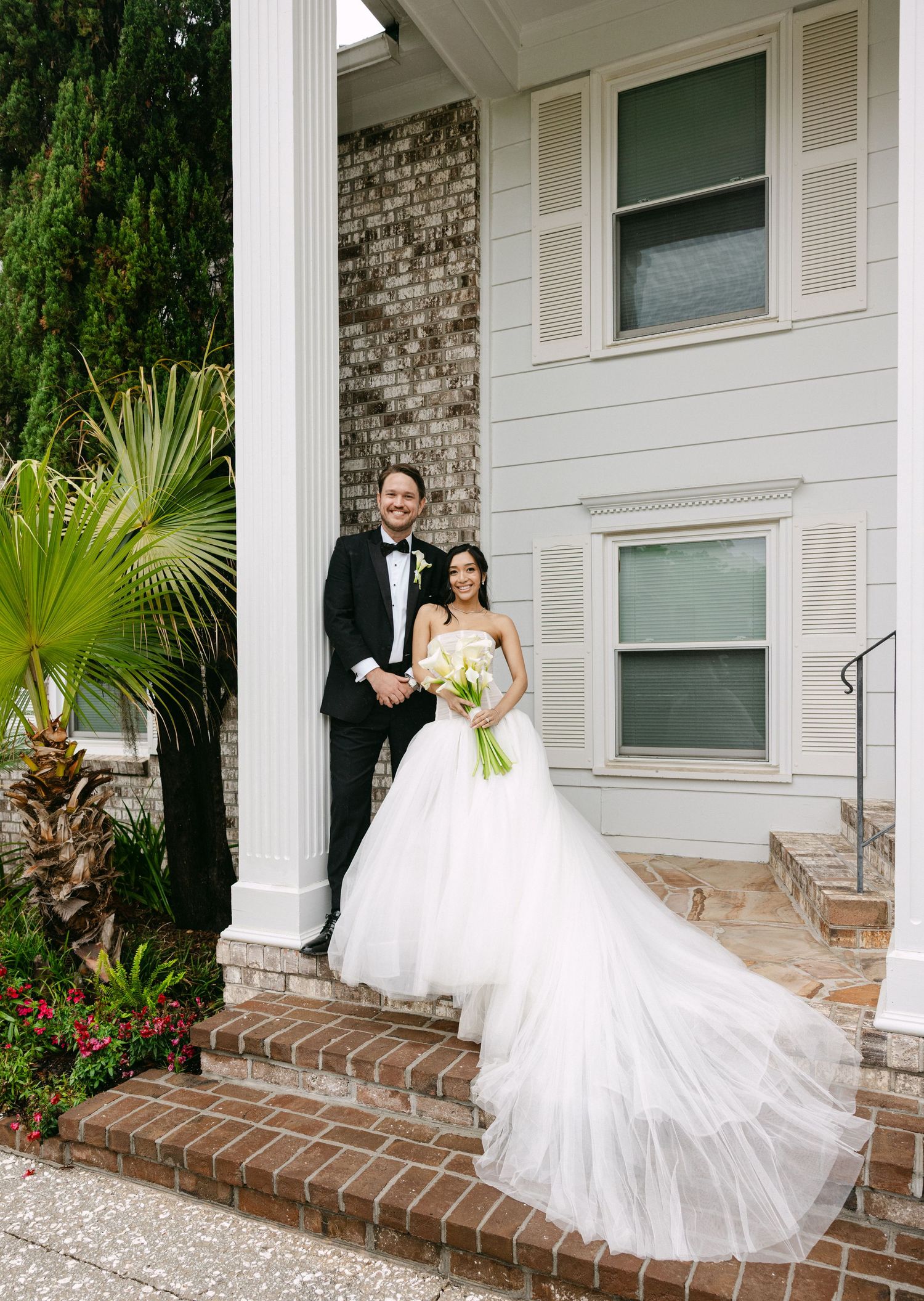 Bride in flowing white gown and groom in black tuxedo pose on brick steps of elegant venue with white columns.