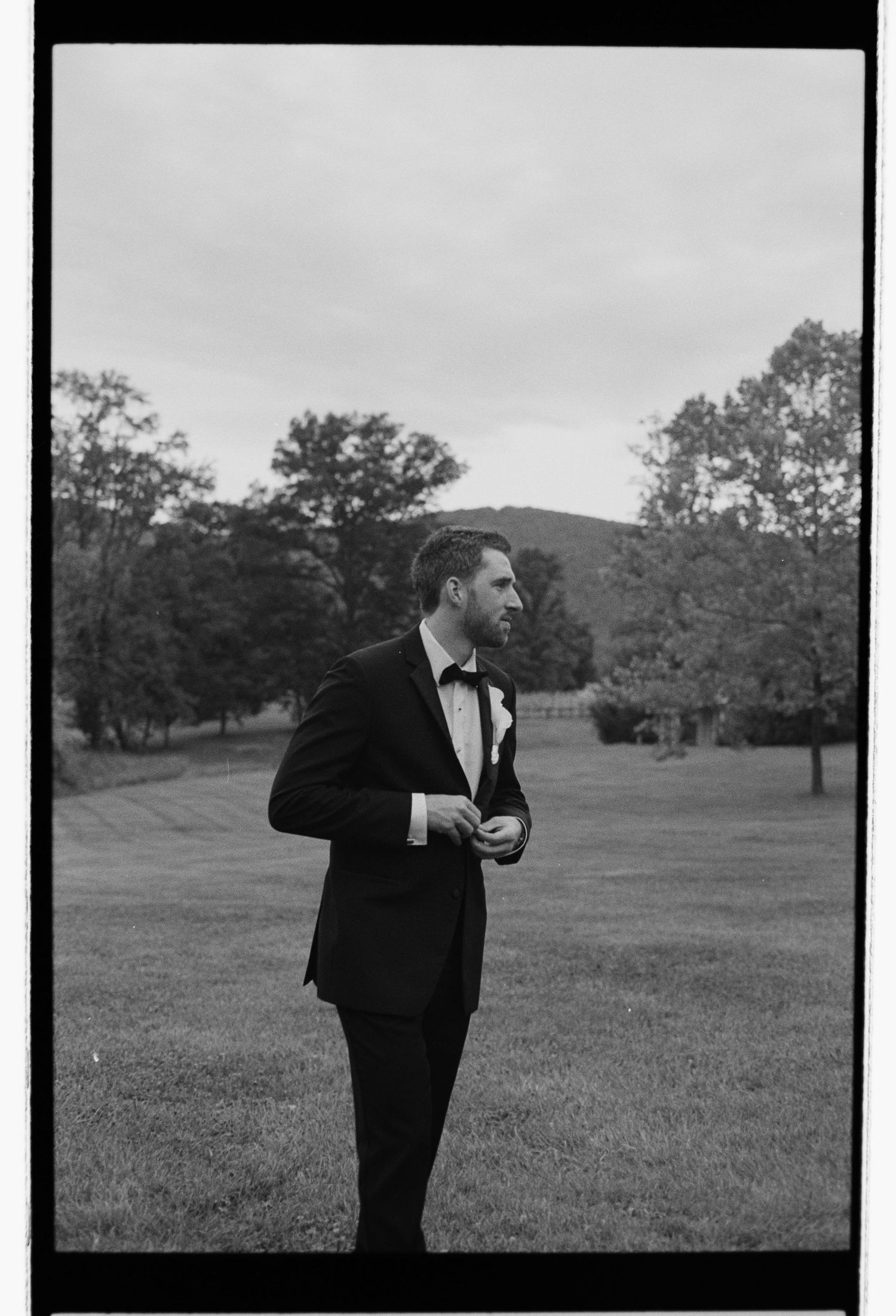 Black and white portrait of groom in tuxedo standing on grassy field with trees and hills in background.