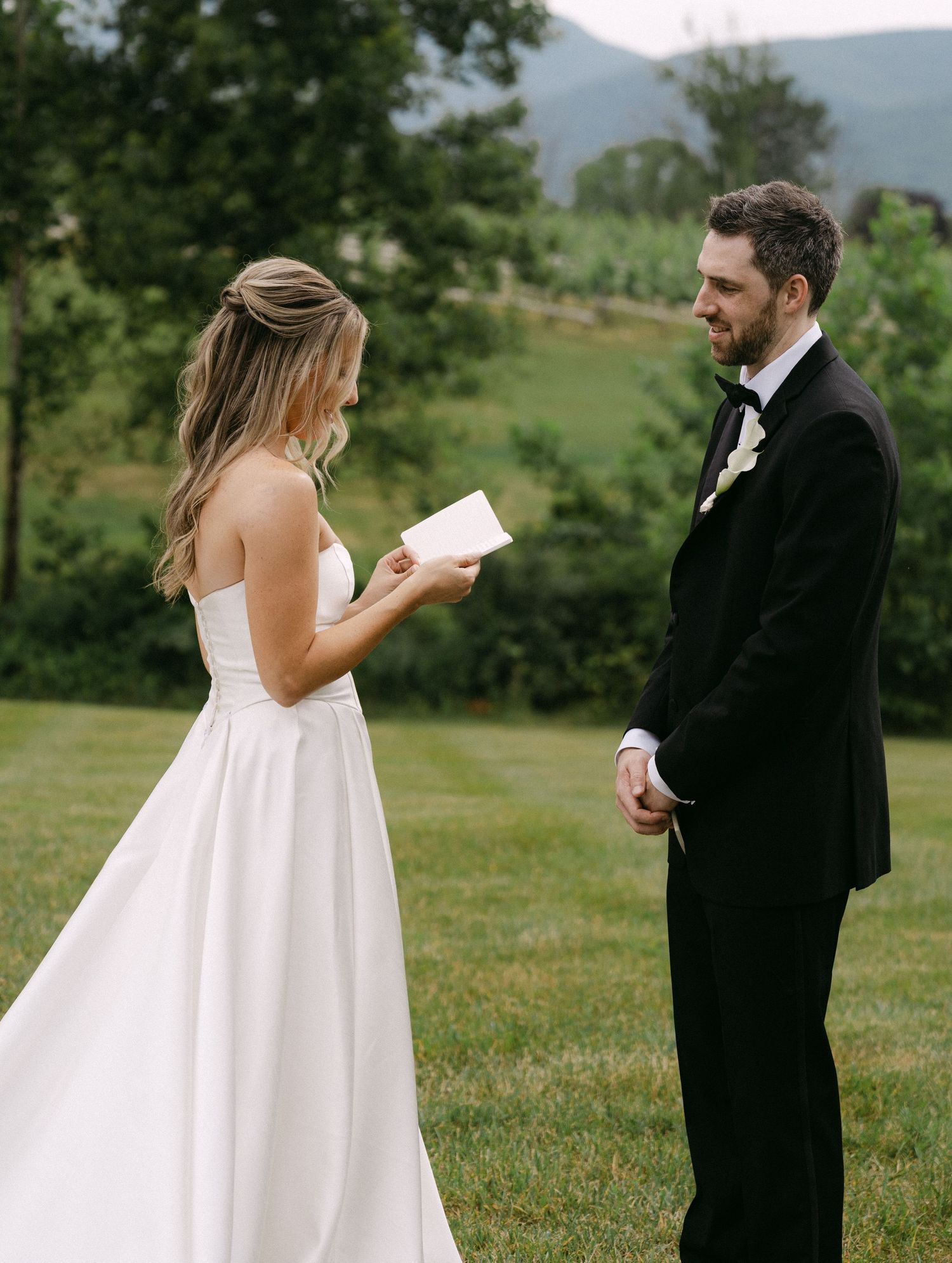 Bride reads vows from a card to groom in black tuxedo during outdoor wedding ceremony with green hills backdrop.