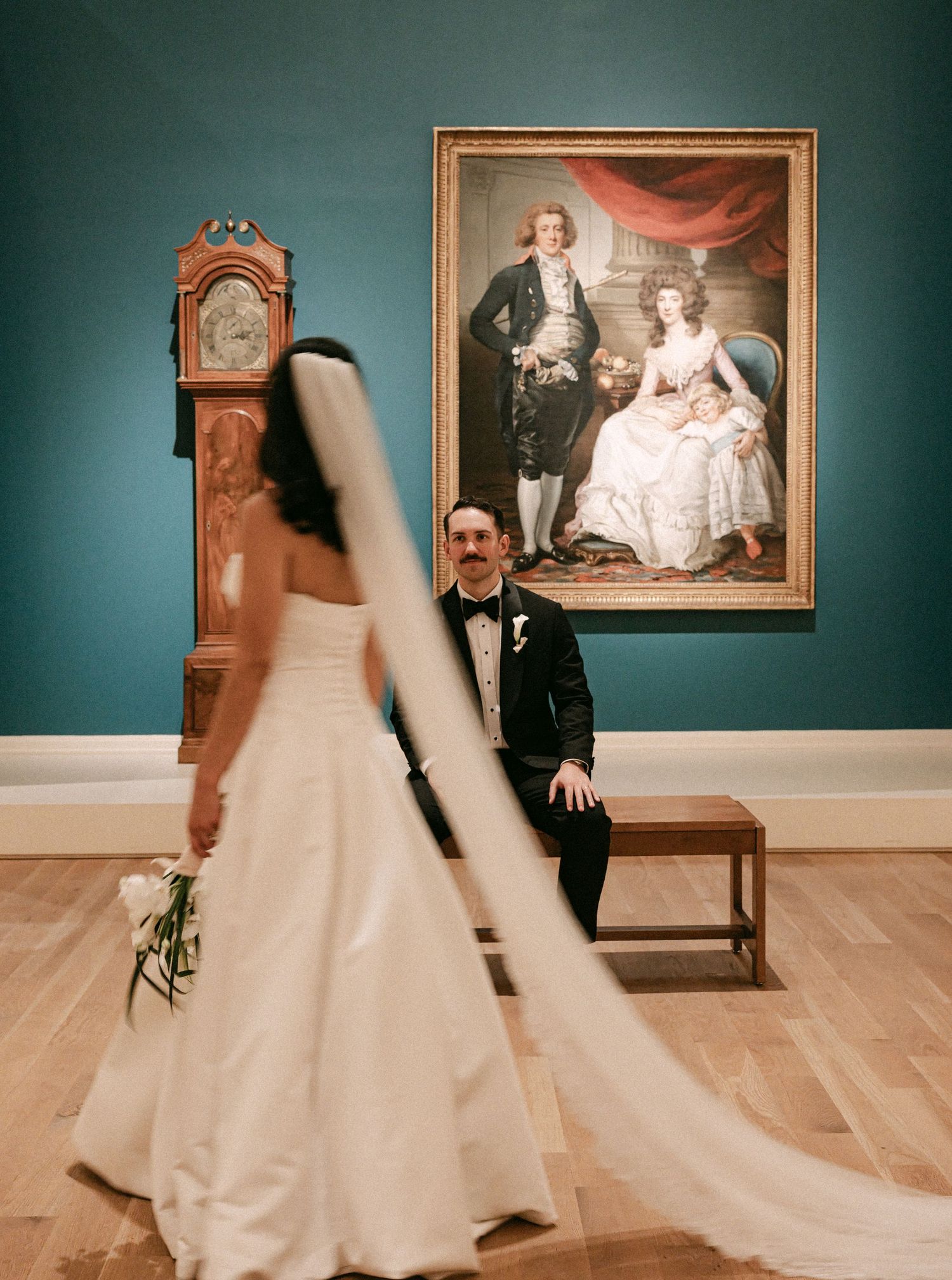 Groom in tuxedo sits smiling as bride in white gown with veil walks toward him before portrait painting.