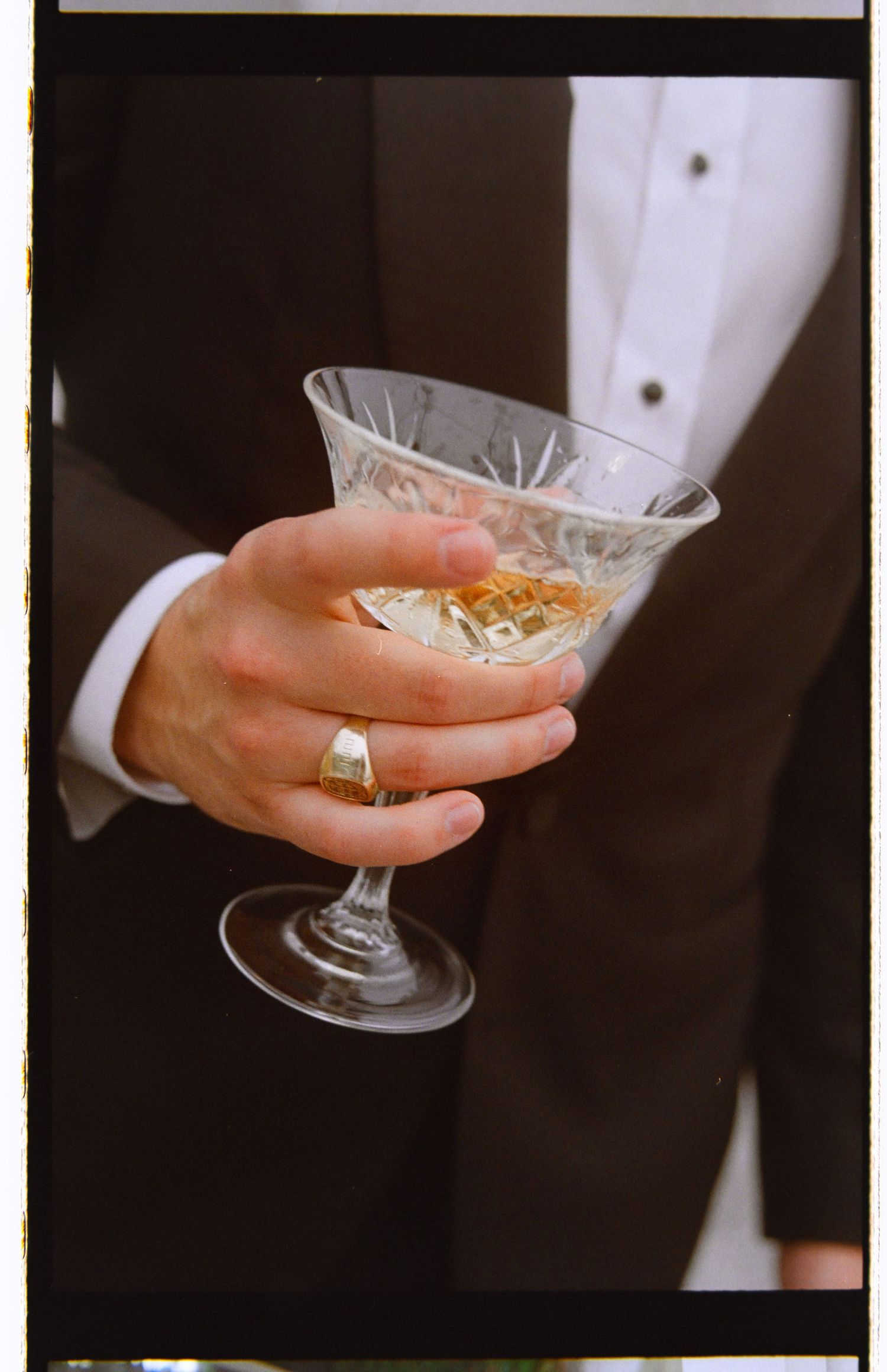 Close-up of groom's hand holding crystal cocktail glass wearing a gold signet ring at wedding reception.