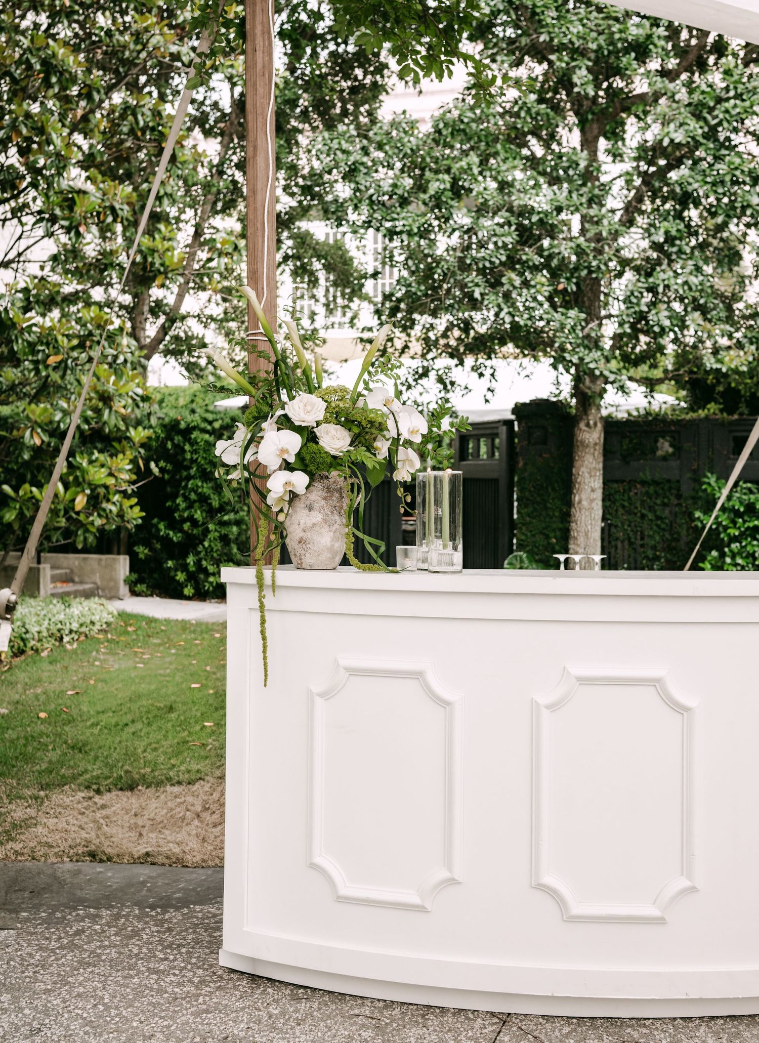 Elegant white curved bar with floral arrangement and lush greenery backdrop at an outdoor wedding reception.