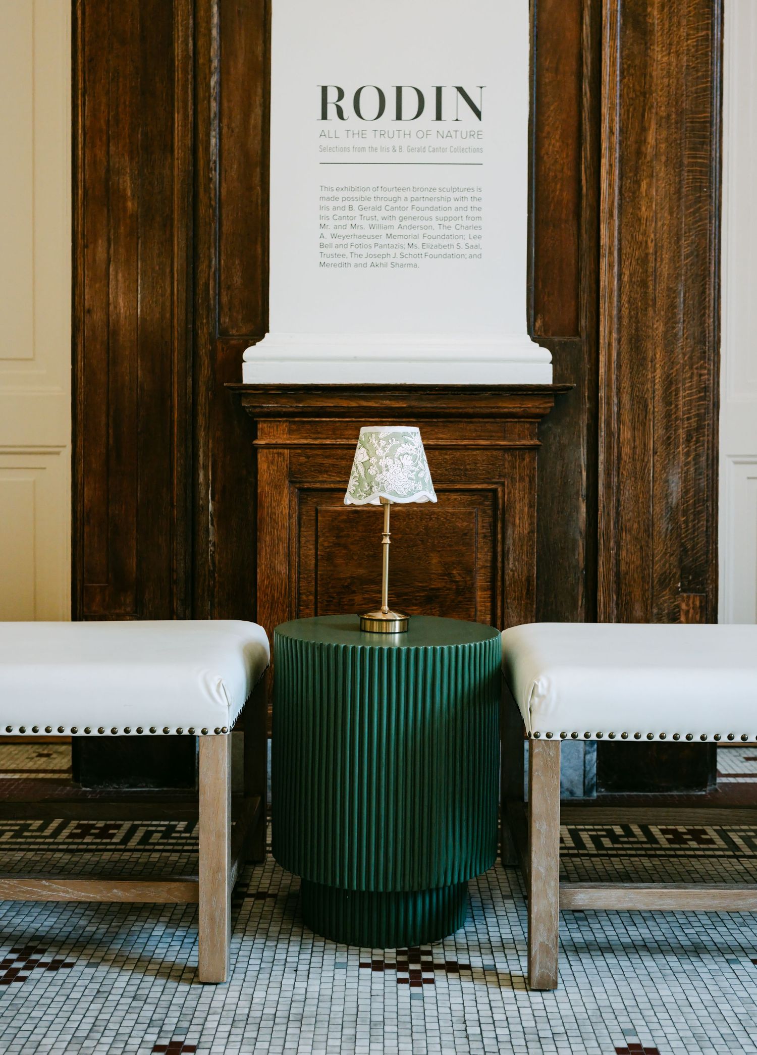 Elegant museum lobby with green ribbed side table, brass lamp, white benches, and Rodin exhibition signage on wood-paneled wall.
