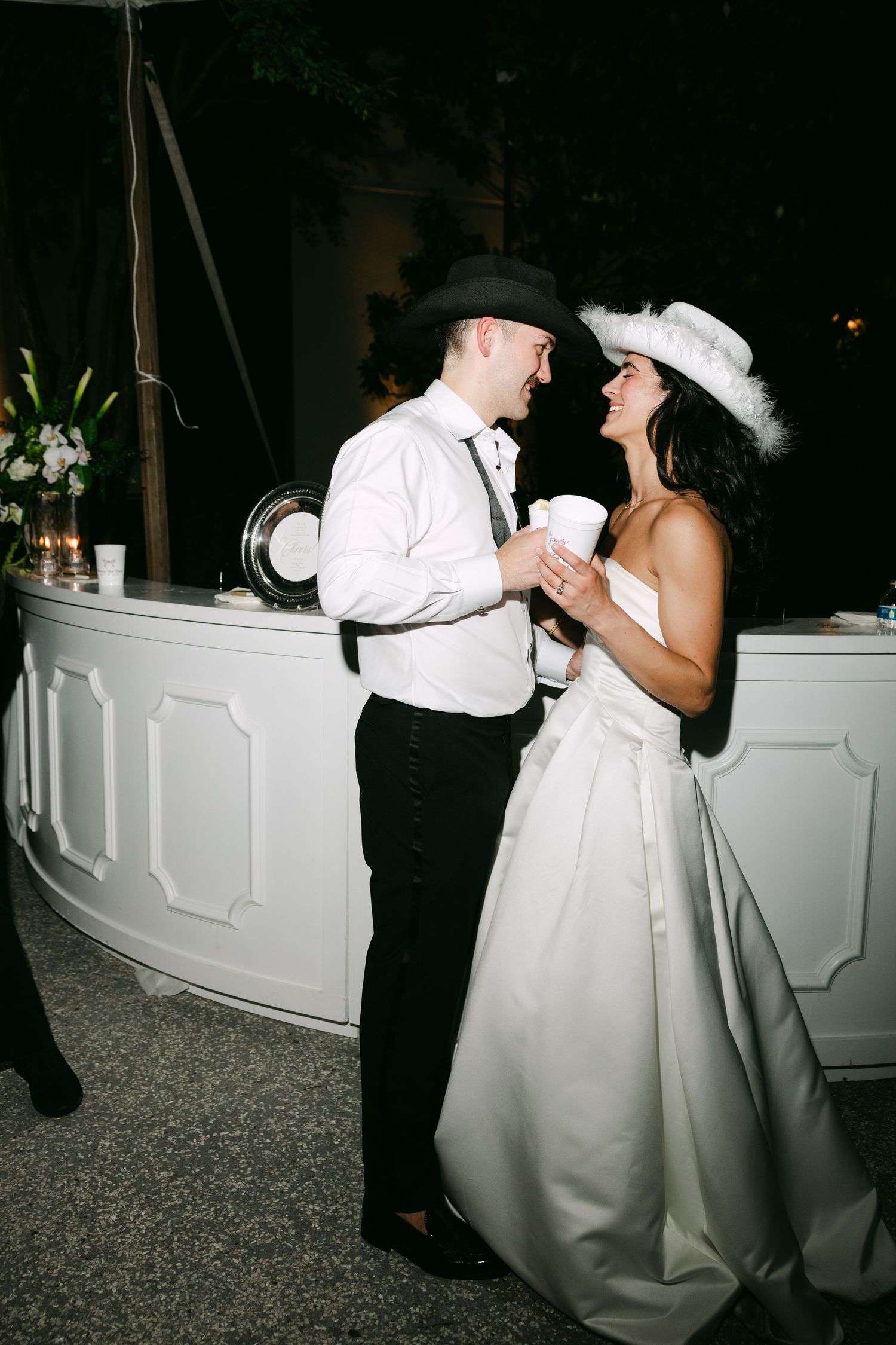 Bride and groom in cowboy hats laugh together at outdoor wedding reception bar.