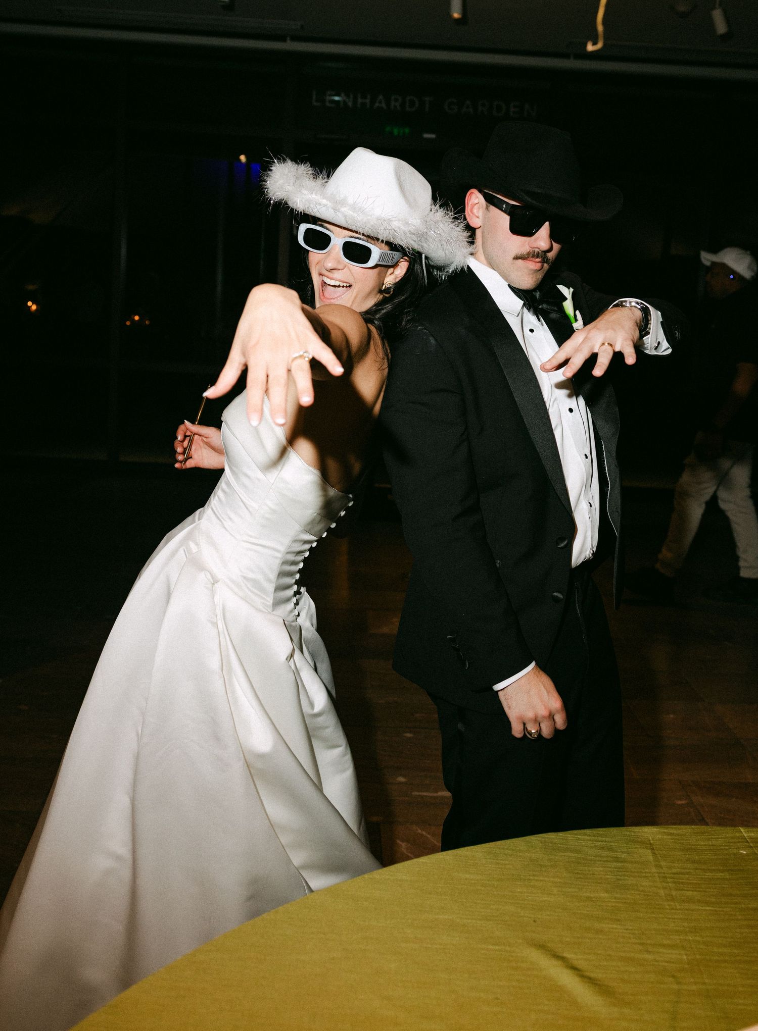 Bride and groom dancing in cowboy hats and white sunglasses at a lively wedding reception.