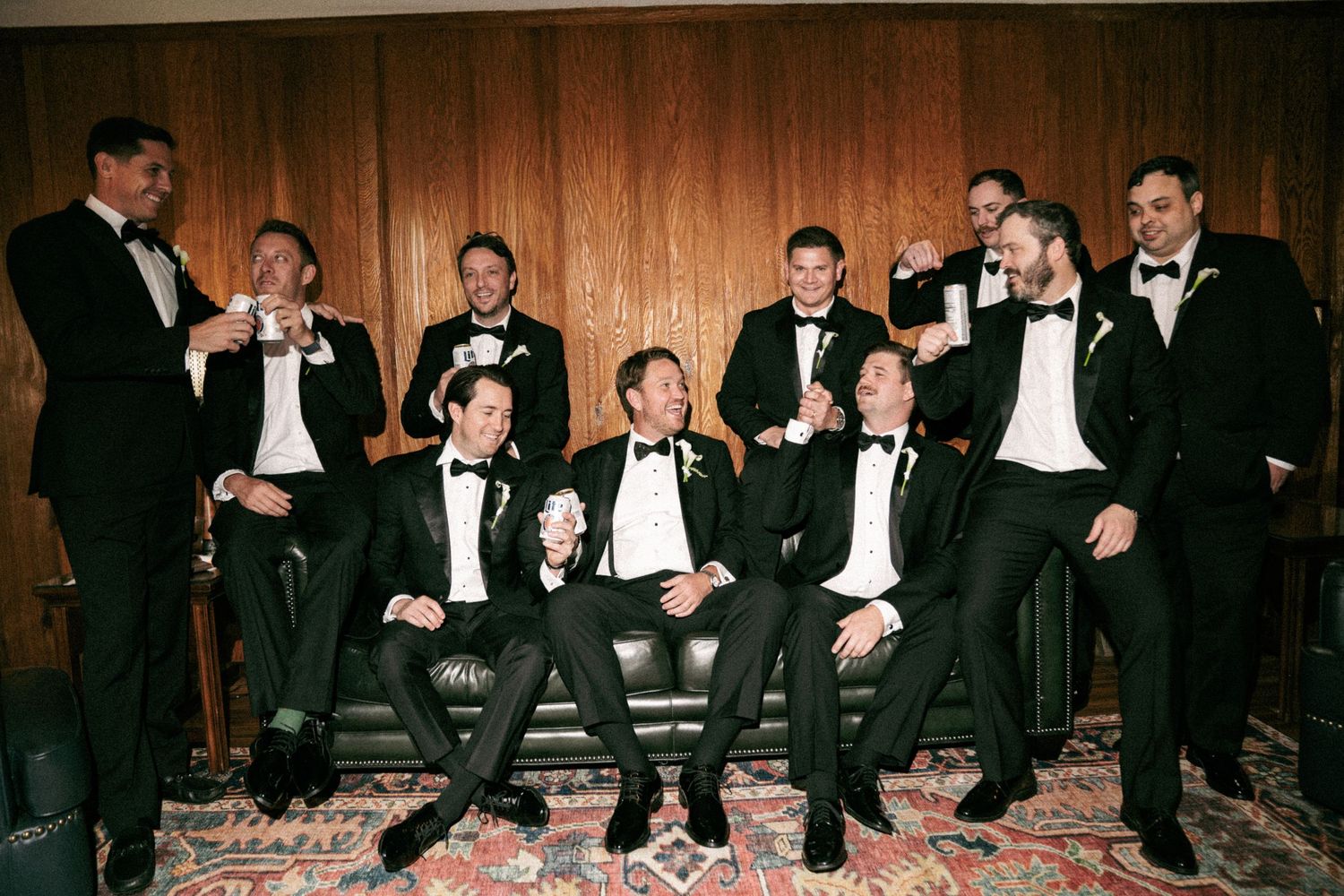 Groomsmen in black tuxedos laughing and toasting drinks together in an elegant wood-paneled room.
