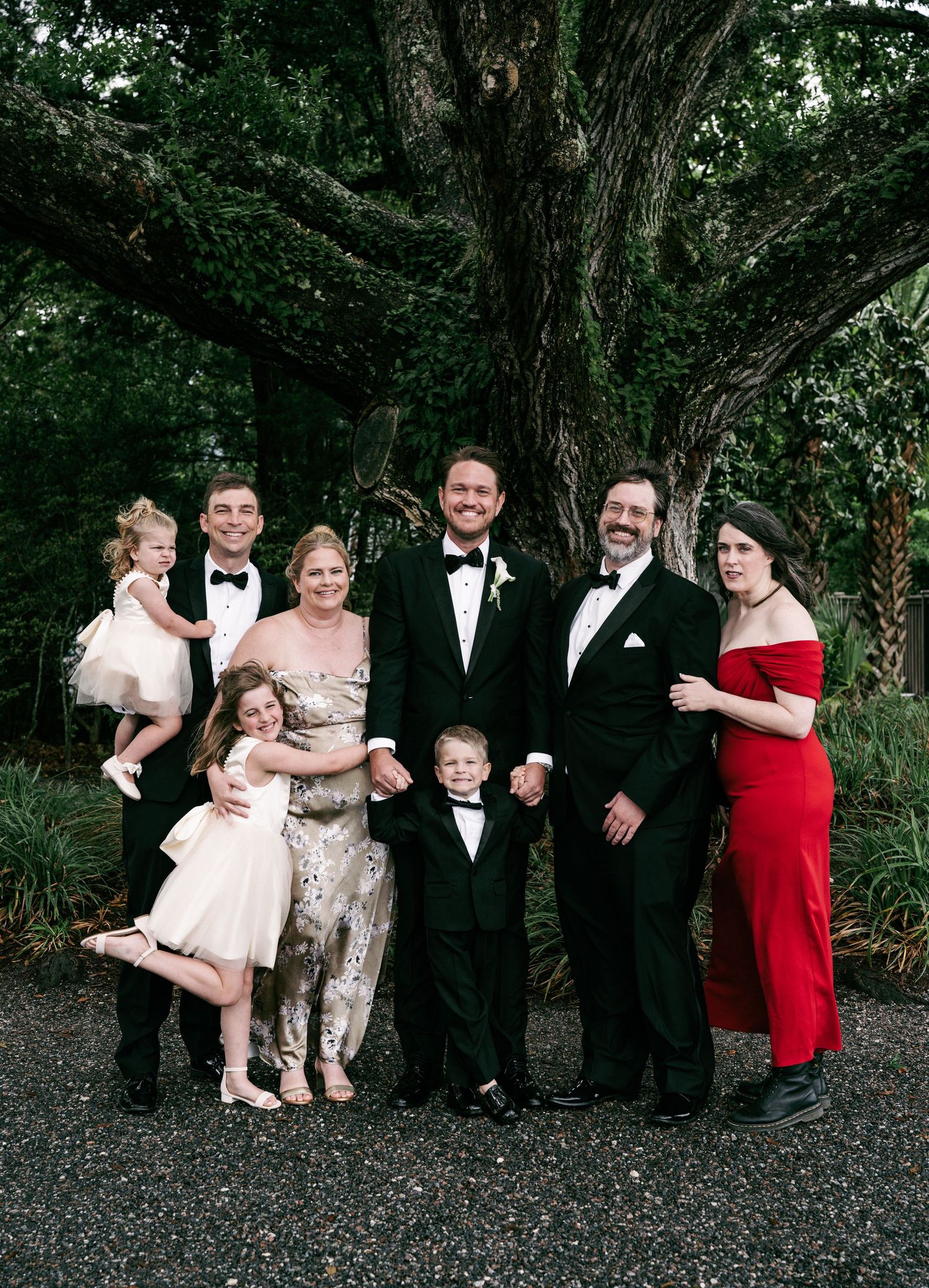 Wedding party family portrait with groom and relatives in formal attire under large oak tree.
