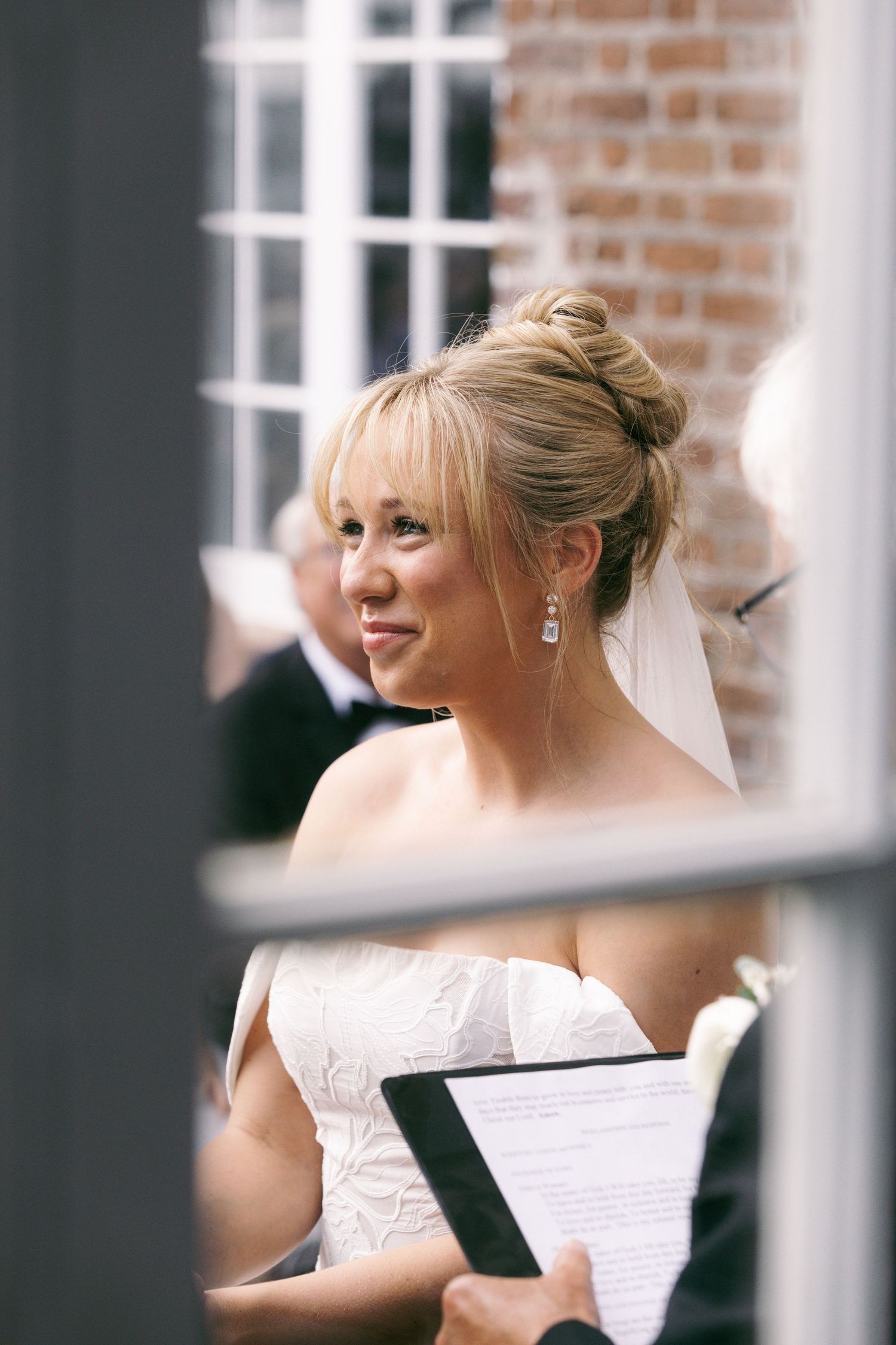 Bride in white lace dress and veil holds clipboard during outdoor wedding ceremony near brick wall.