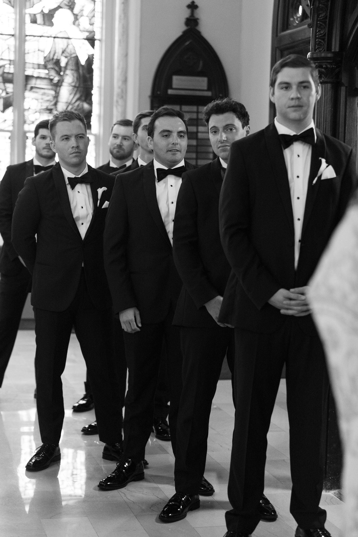 Groomsmen in black tuxedos pose for formal wedding photos in church hallway.