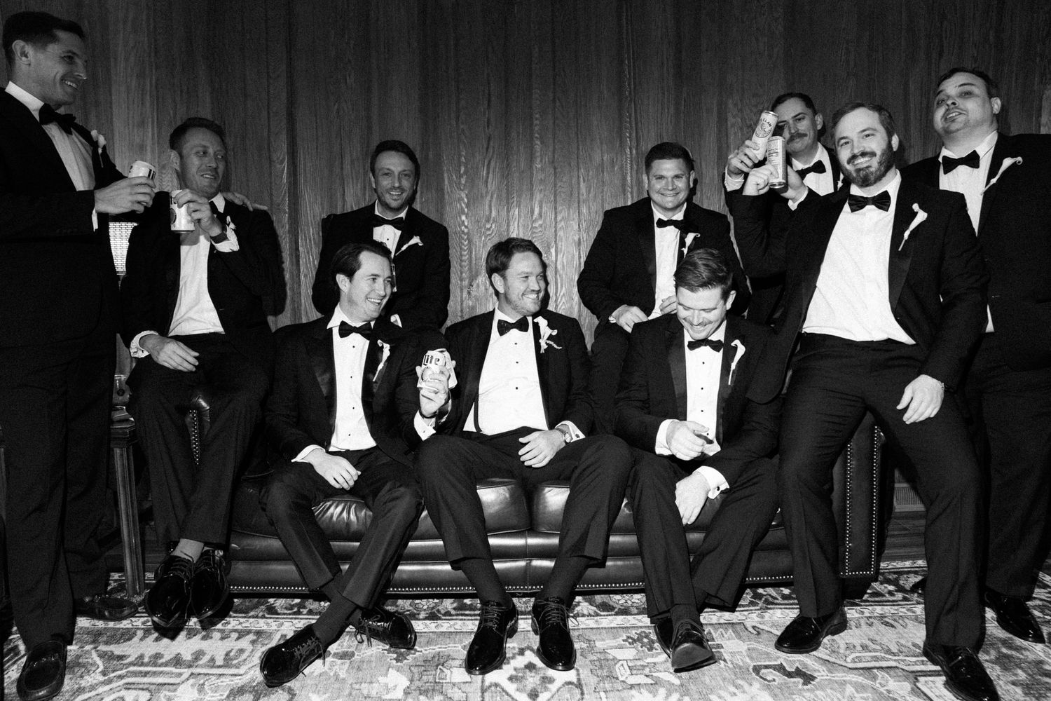 Black and white photo of groomsmen in tuxedos laughing and posing together at a wedding celebration.