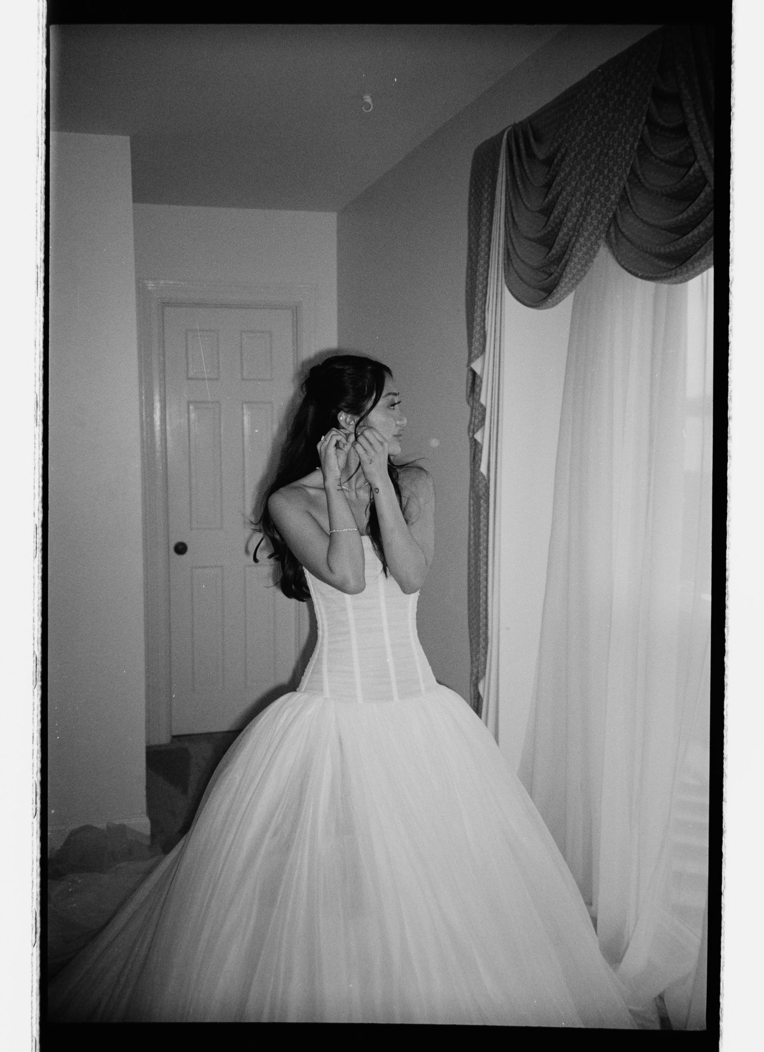 Black and white photo of a bride in a strapless ballgown adjusting her earring by a curtained window.