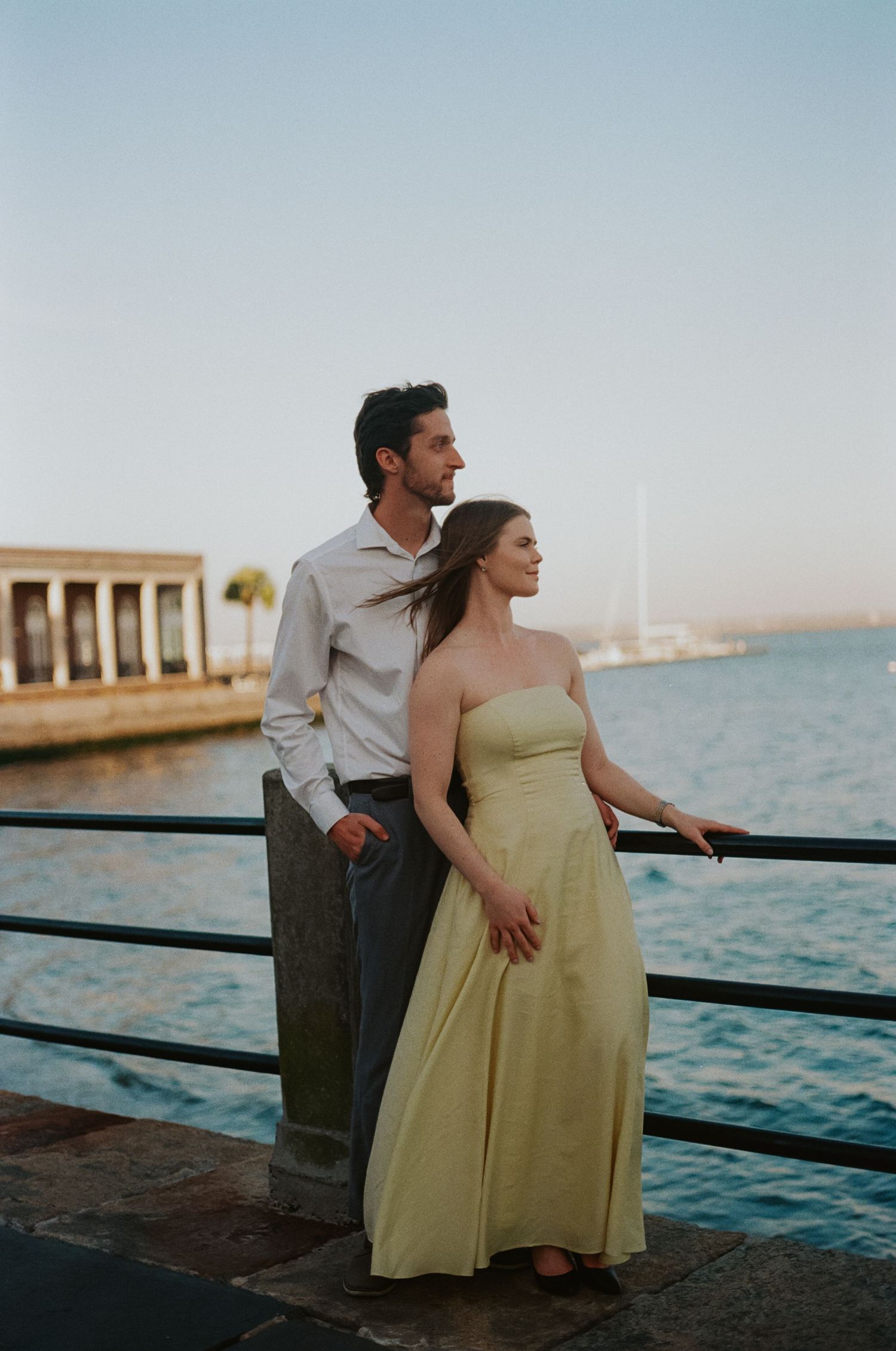 Couple standing by waterfront railing at sunset, woman in yellow strapless gown, man in white shirt, gazing at the sea.