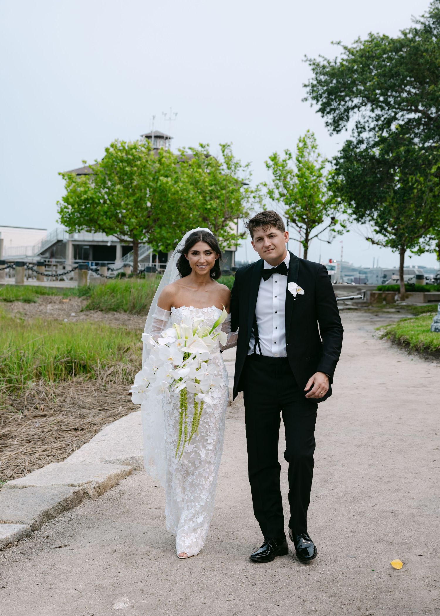 Wedding couple poses along a winding dirt path with trees and urban landscape in background.