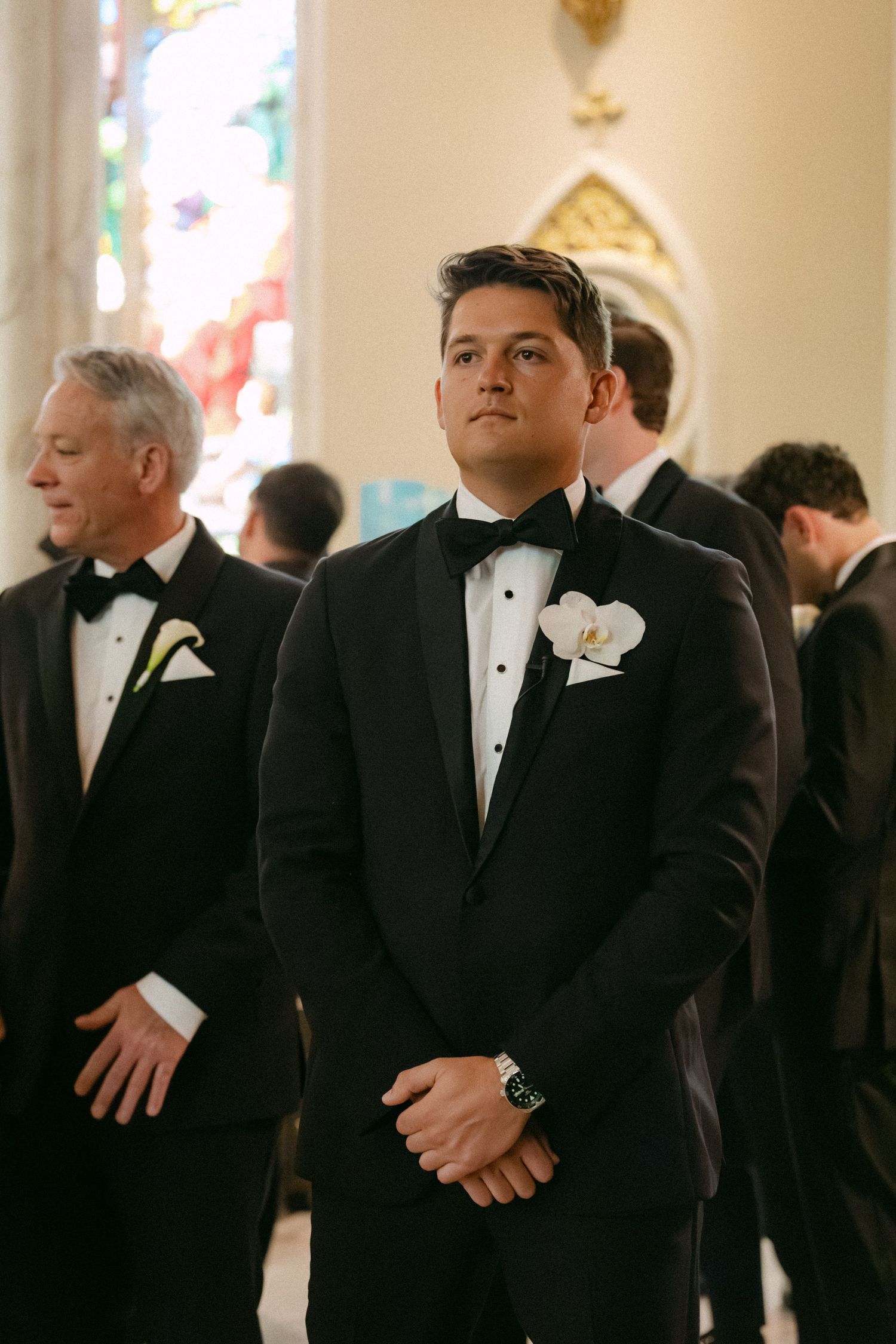 Groomsmen in black tuxedos stand in formation inside a church with stained glass windows.