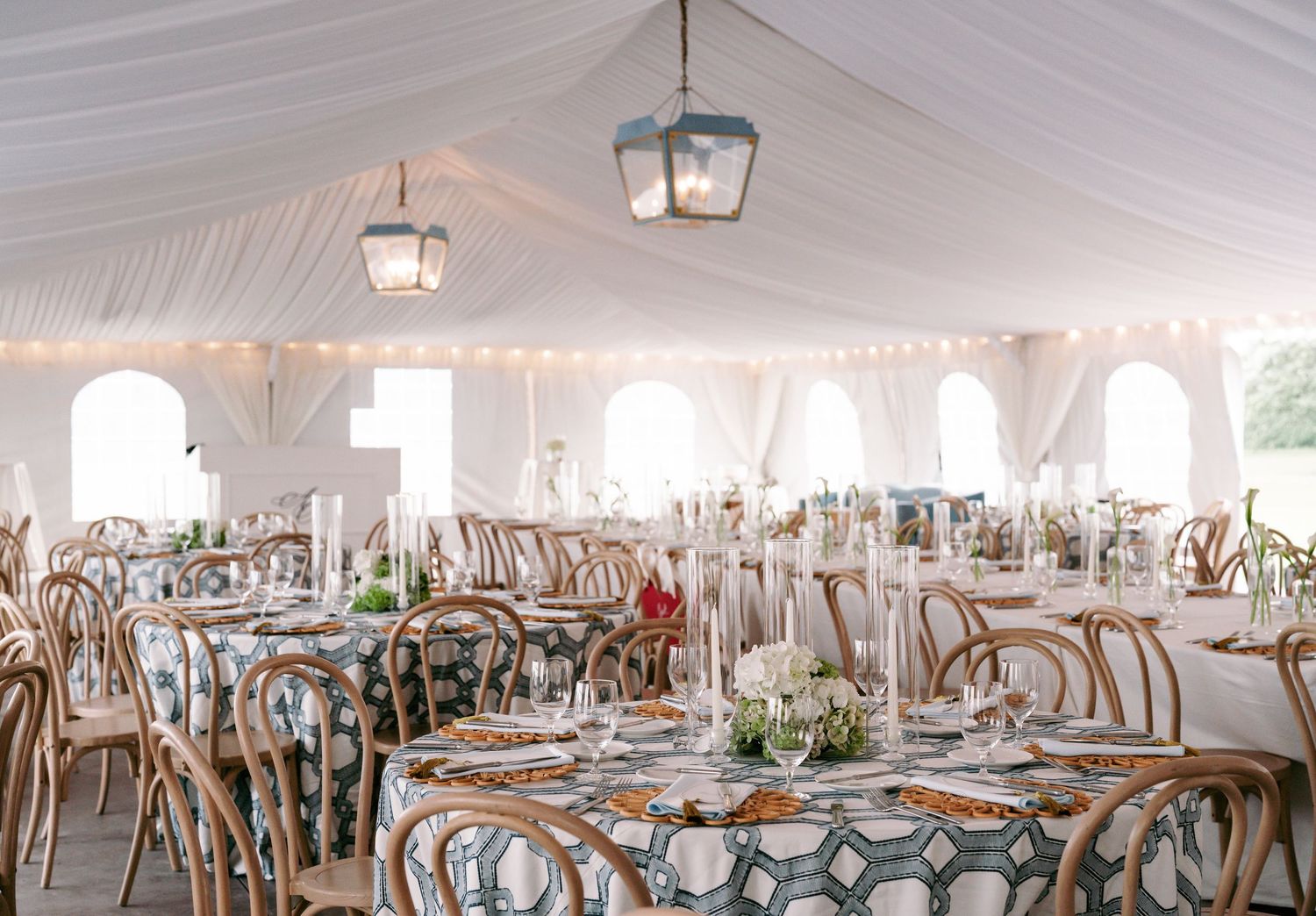 Bright wedding reception tent with bentwood chairs, patterned tablecloths, hanging lanterns, and arched windows.