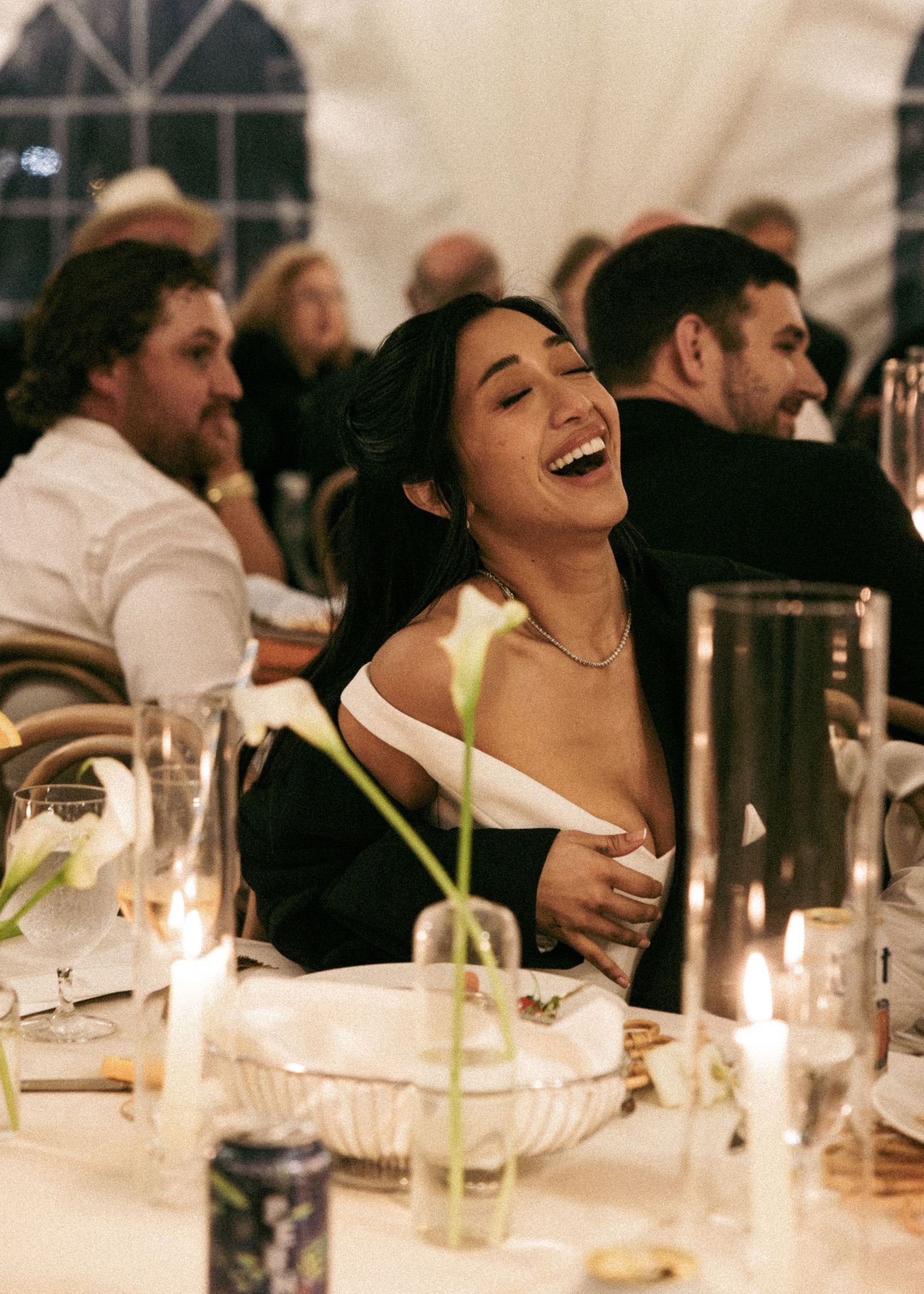 A joyful wedding guest in an off-shoulder black and white outfit laughing at a candlelit reception dinner table.