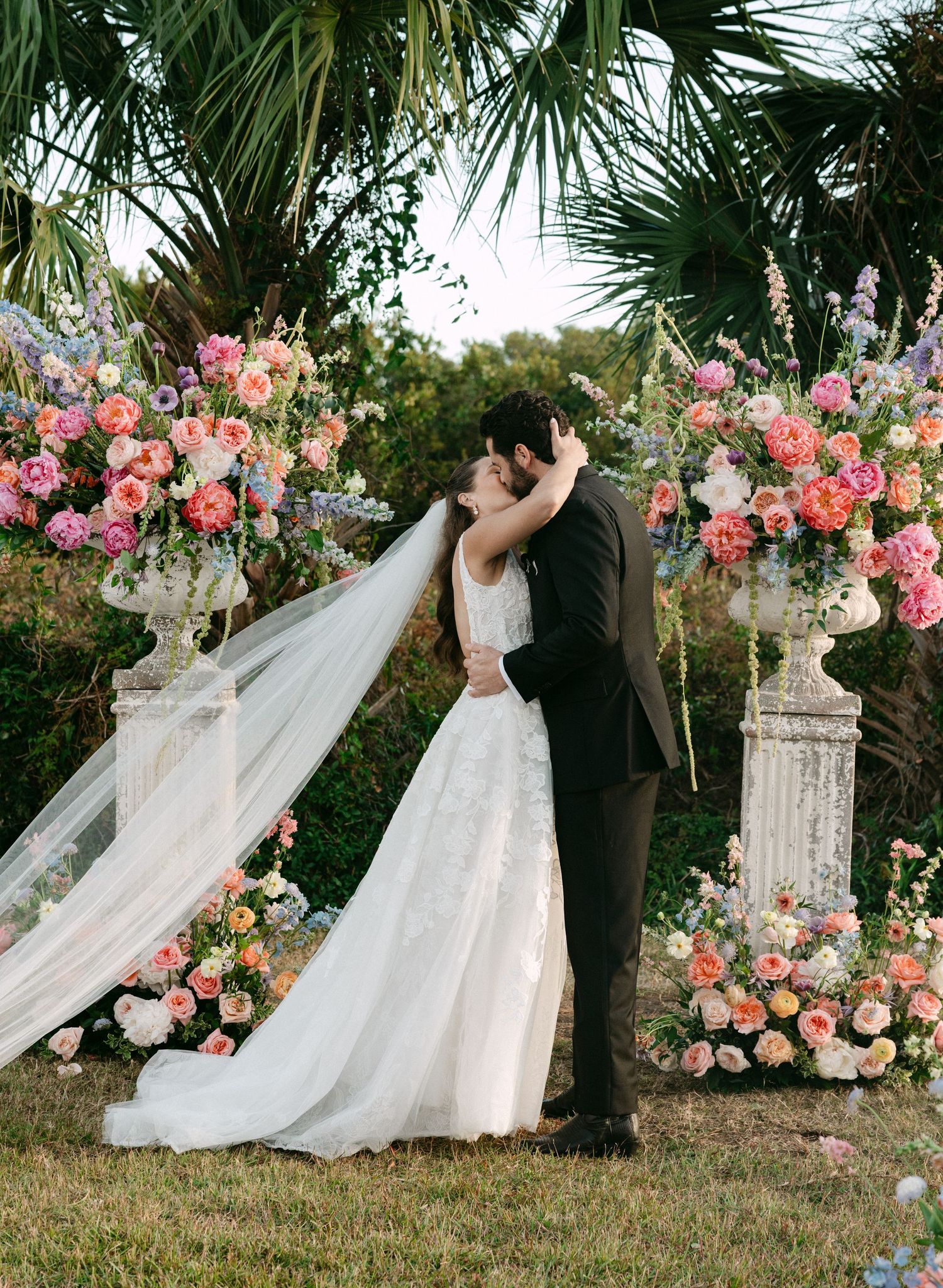 Bride and groom share first kiss at floral altar with pink and coral blooms at outdoor wedding.