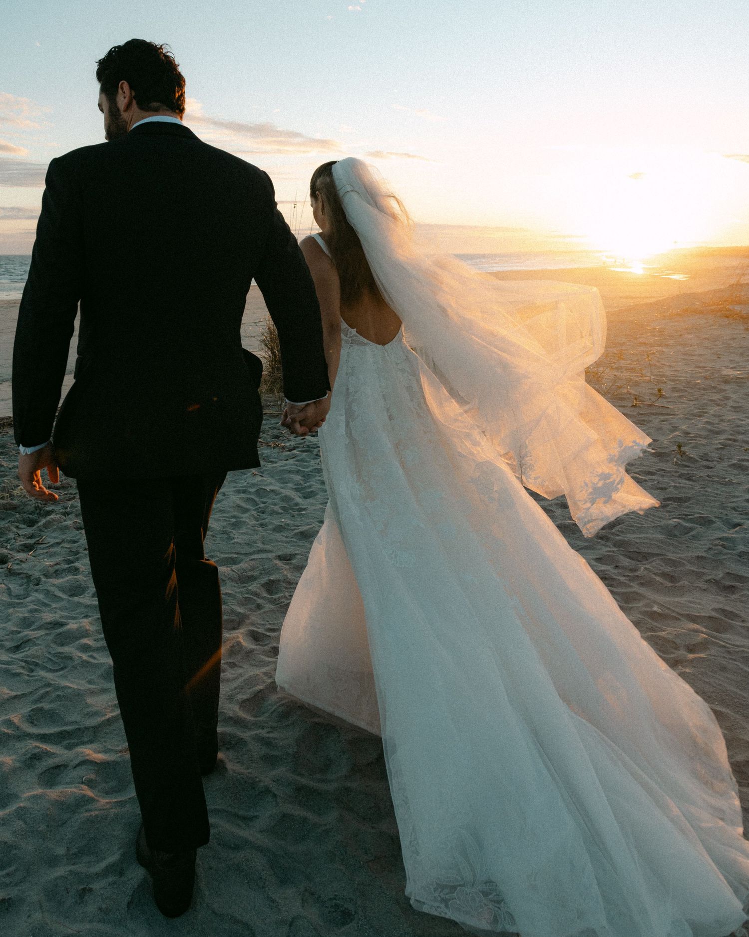Bride and groom walk hand-in-hand on a sandy beach at sunset in wedding attire.