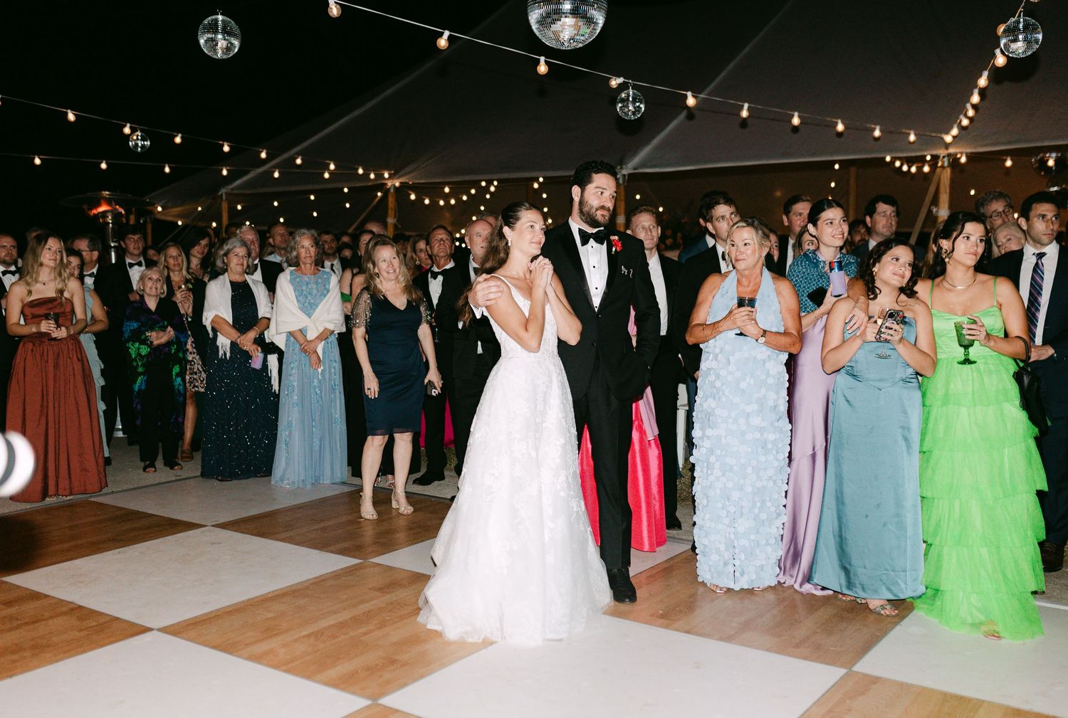 Bride and groom share first dance on checkered floor under string lights at wedding reception.