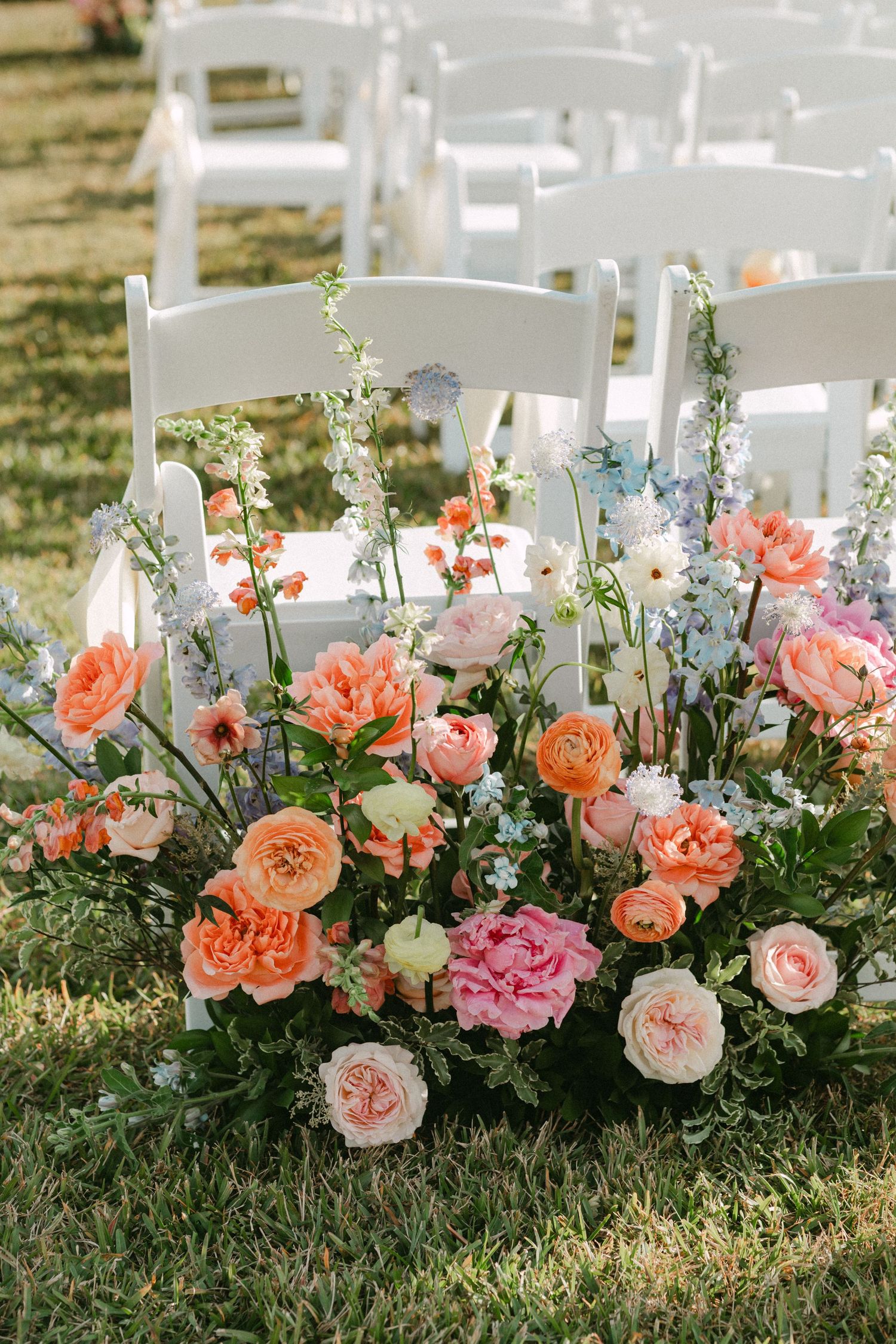 Lush arrangement of peach, pink, and blue flowers decorating white chairs at outdoor wedding.