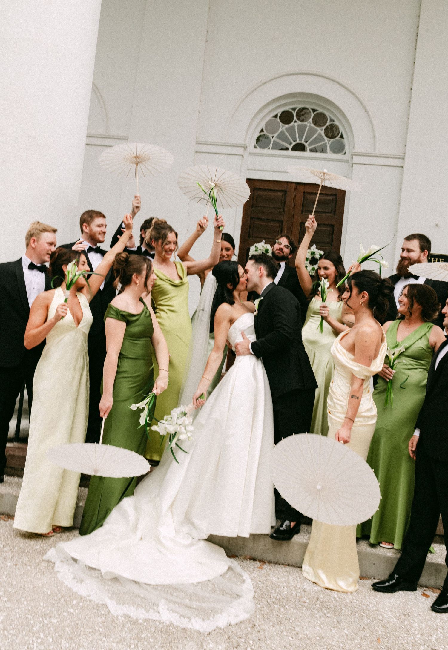 Wedding party celebrating outside a white church, bridesmaids in green gowns holding parasols as couple kisses.