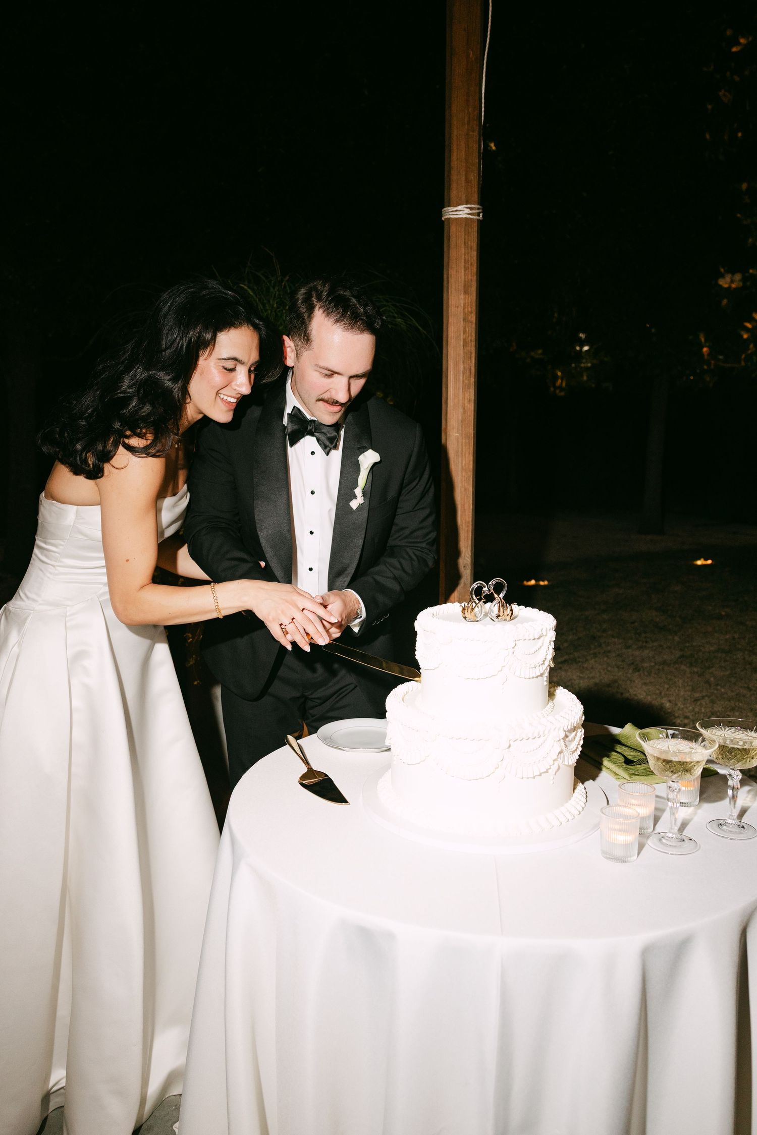 Bride and groom cutting white three-tier wedding cake together outdoors at night.
