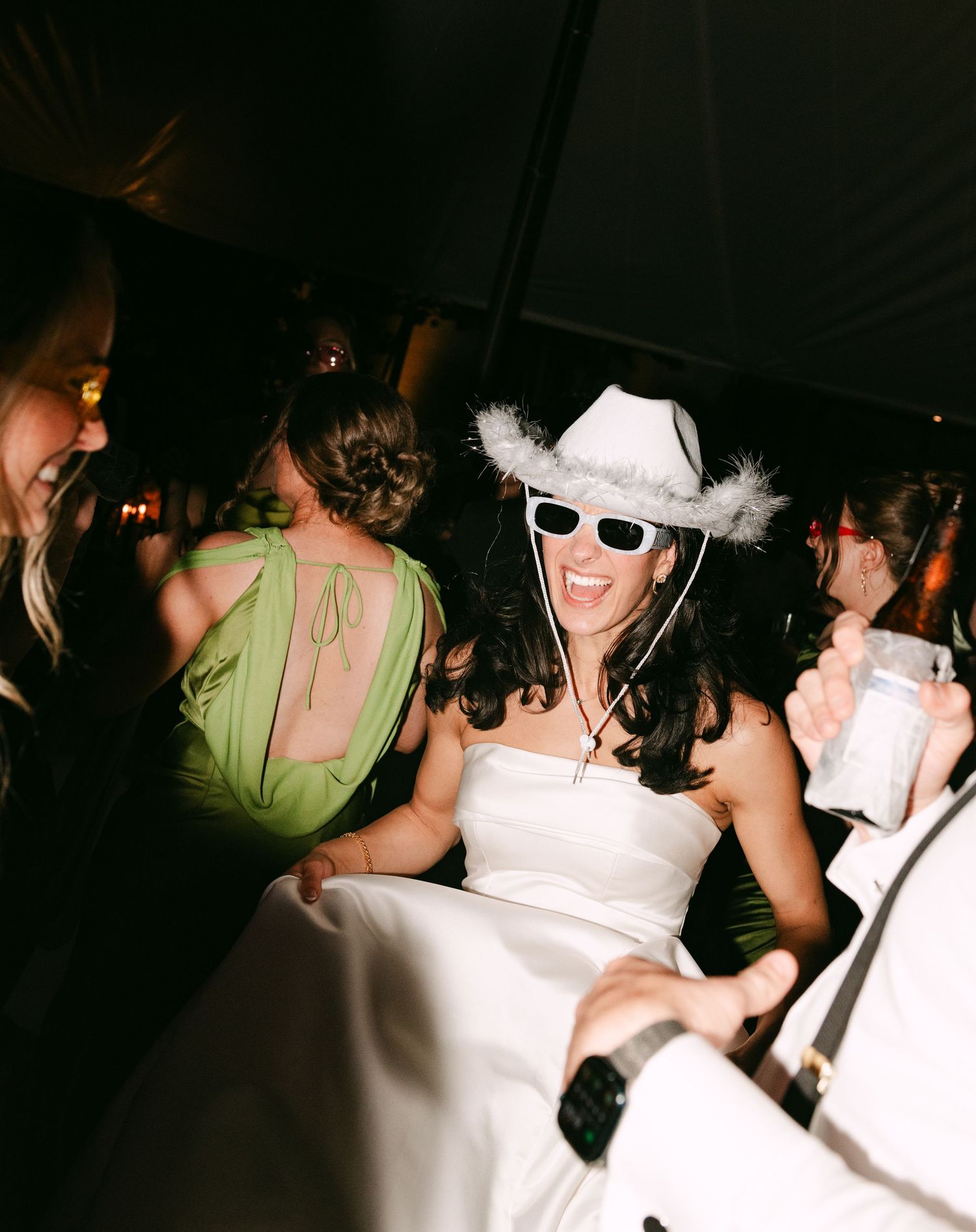 Bride dances joyfully wearing white cowboy hat and sunglasses at lively wedding reception party.