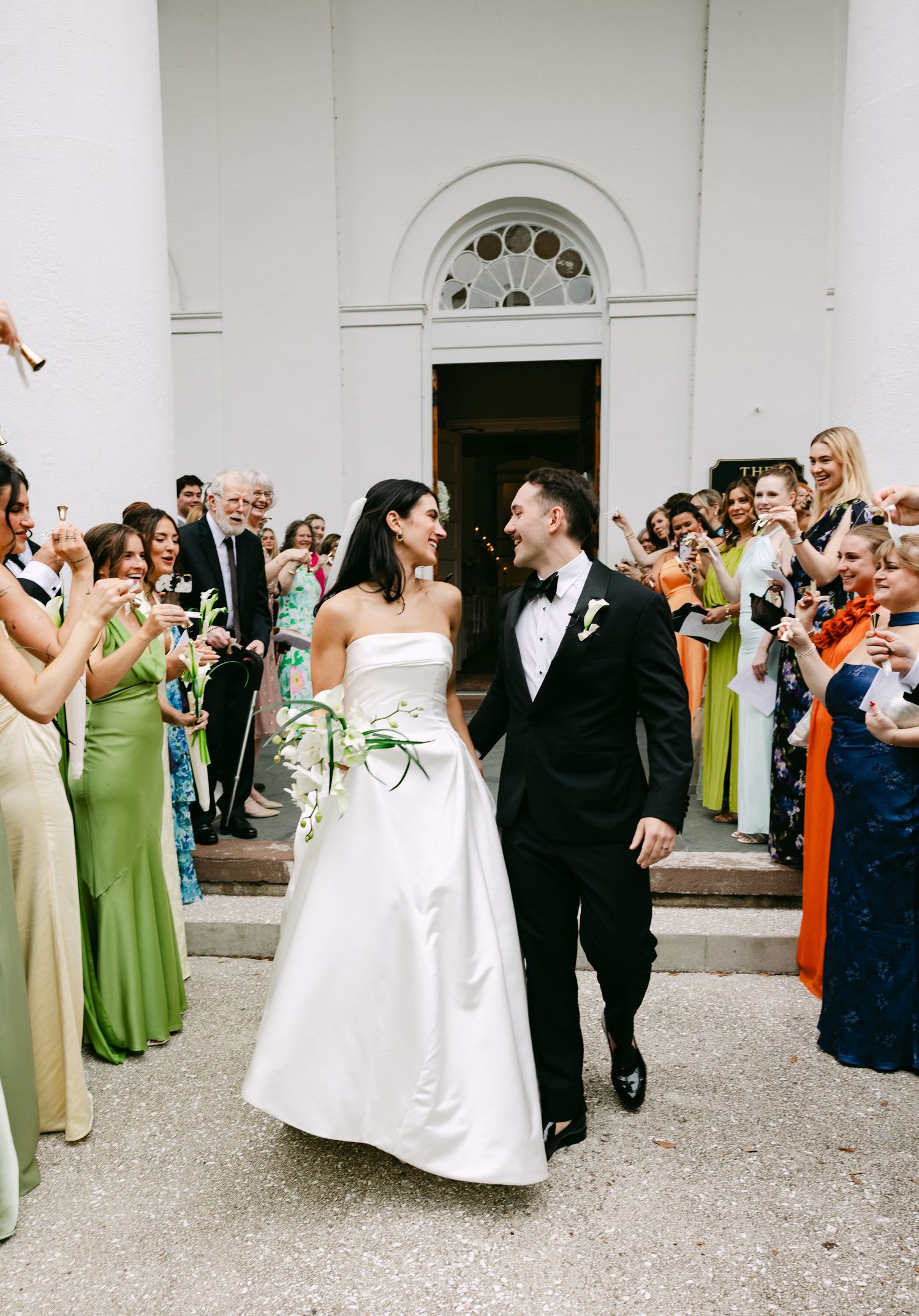 Newlyweds walk through cheering guests outside white church as bride holds tropical bouquet.