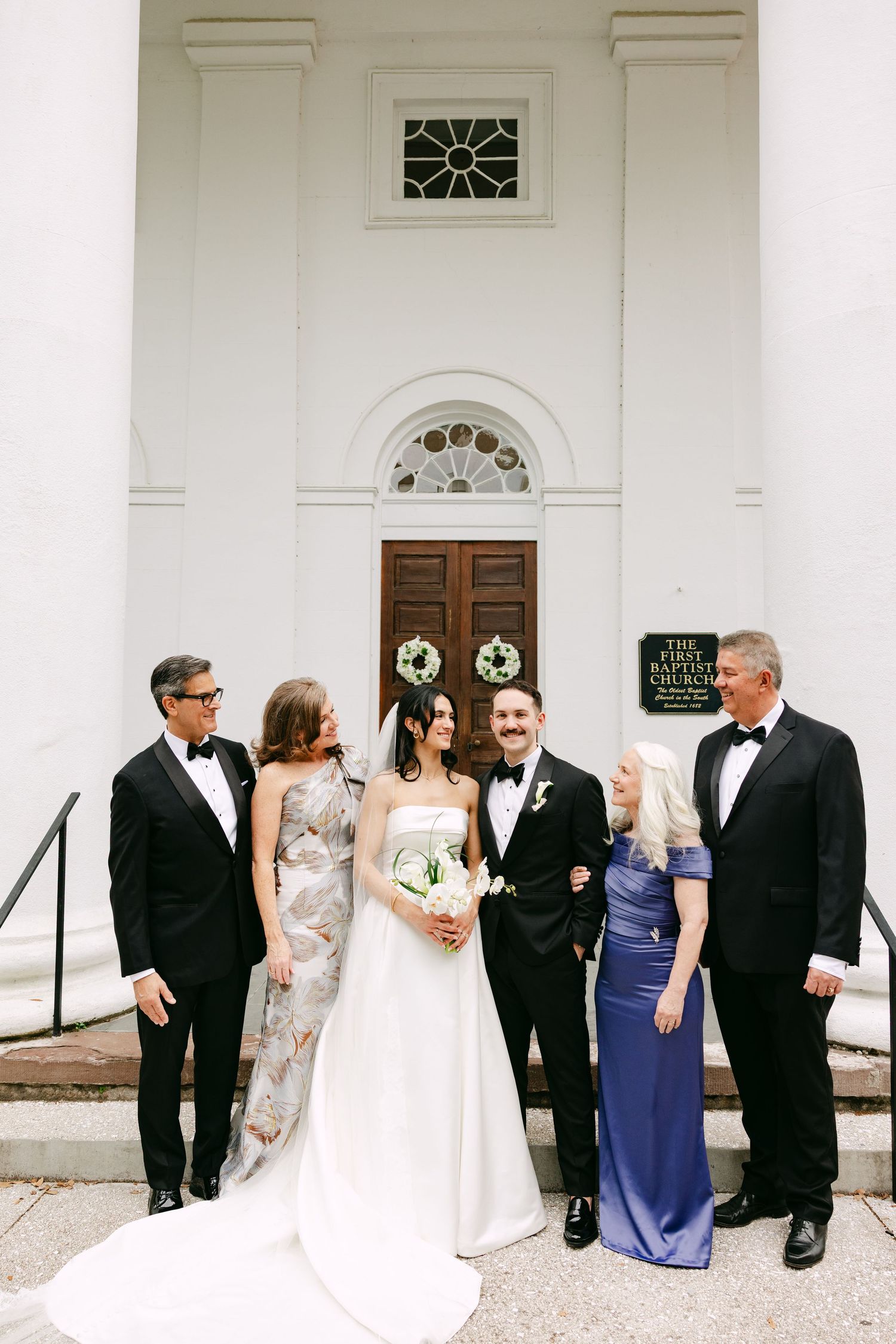Wedding party poses on church steps, bride in strapless gown holding bouquet with family in formal attire.