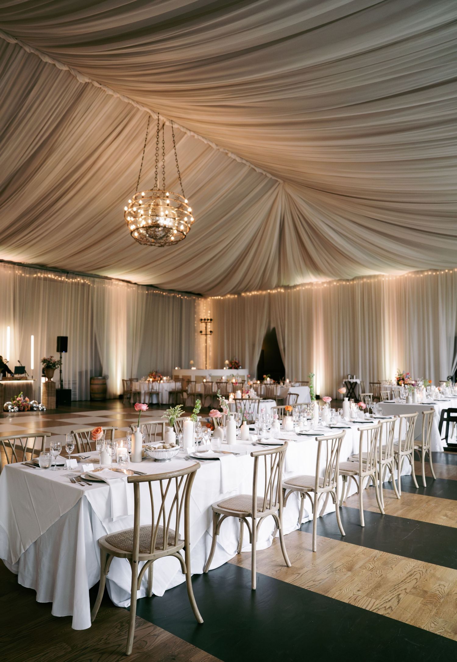 Elegant wedding reception tent with draped white fabric ceiling, chandelier lighting, and long white tablecloths.