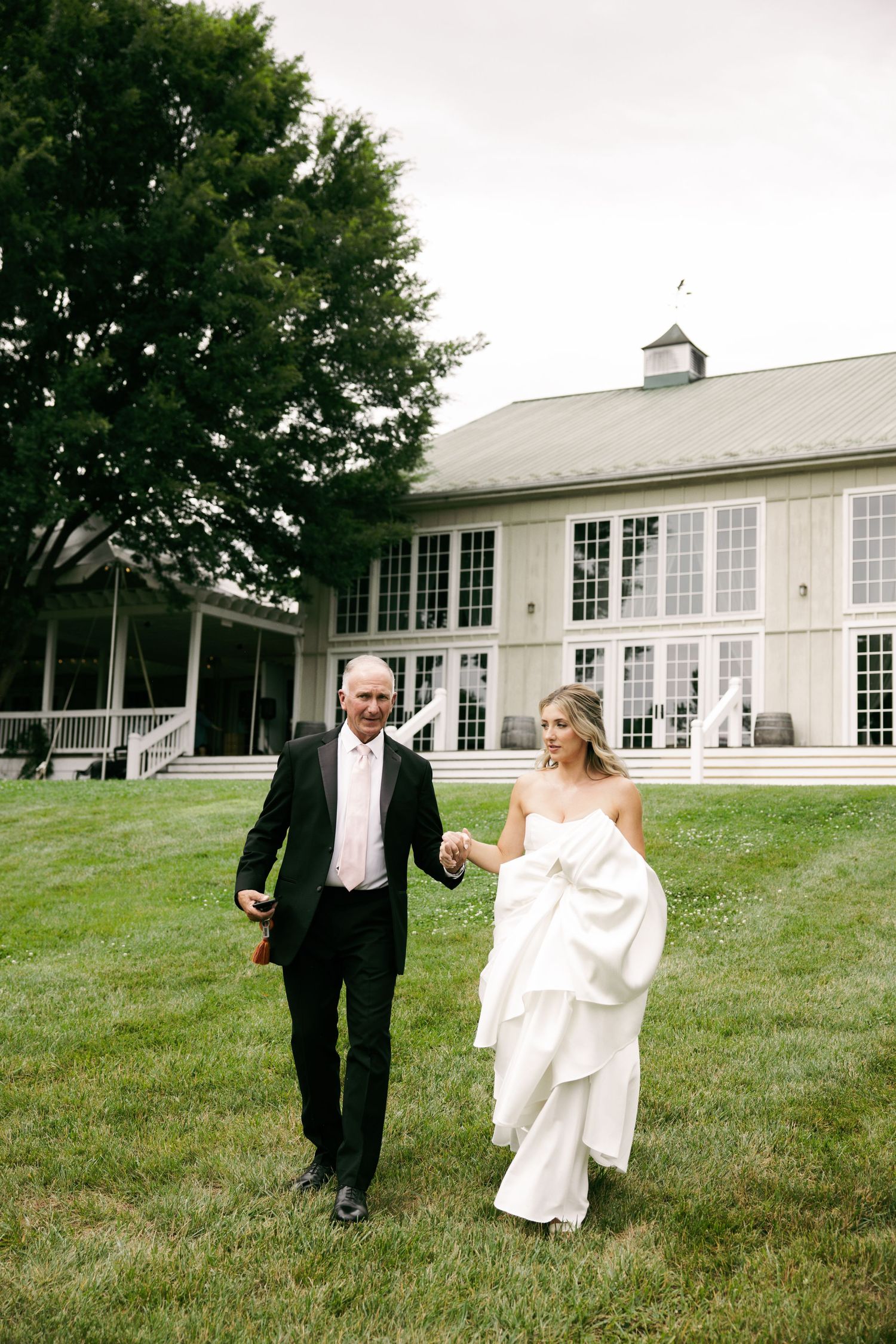 Bride in strapless layered white gown walks hand-in-hand with father across green lawn.