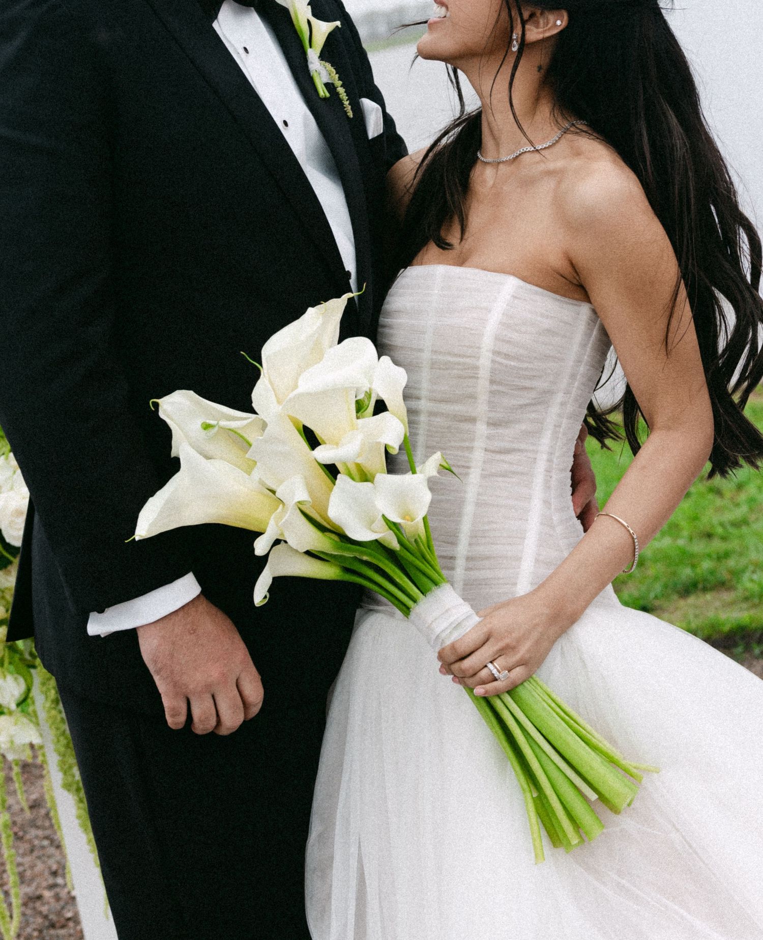 Bride in strapless white gown holding calla lily bouquet stands beside groom in black tuxedo at elegant wedding.