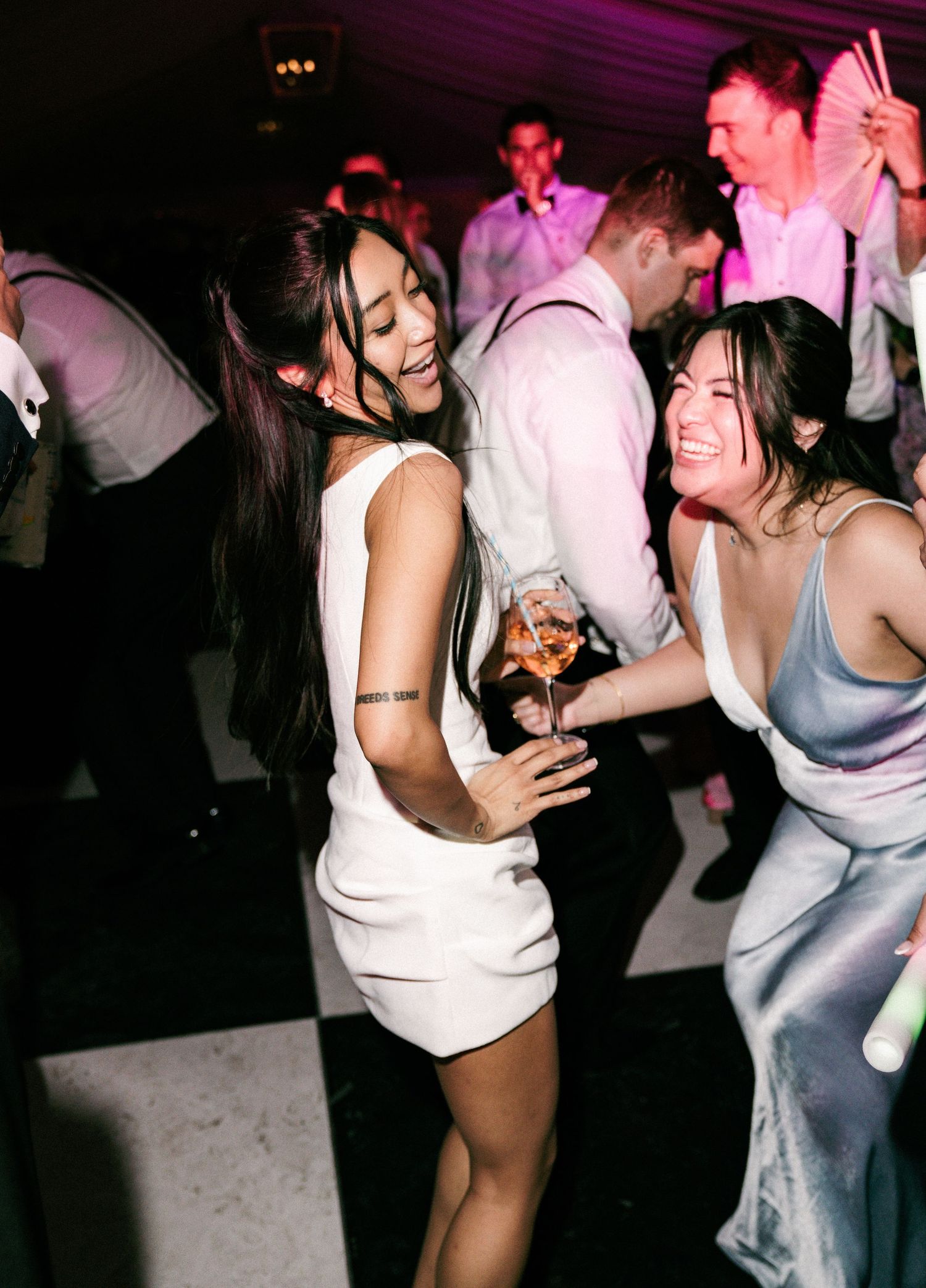 Two women laughing and dancing on checkered dance floor at lively wedding reception party.